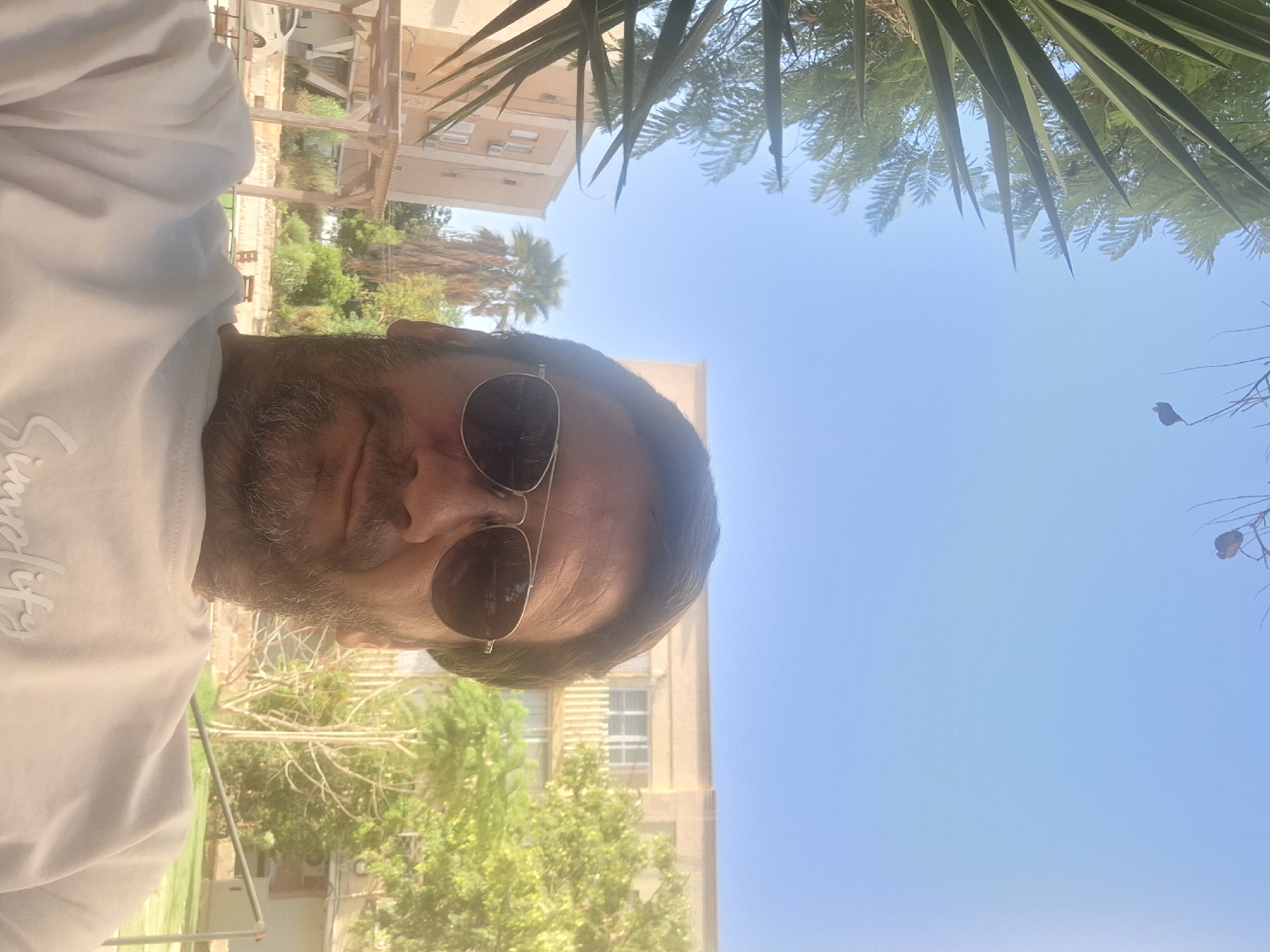 A medium shot captures a man from the chest up, wearing a white t-shirt and aviator sunglasses, as he takes a selfie outdoors. He has short, dark hair and a beard, and is smiling slightly. The background shows buildings, palm trees, and lush green foliage under a clear blue sky, suggesting a warm, sunny day. The man appears to be seated, possibly on a bench or chair. The overall scene suggests a relaxed, casual moment in a residential or garden area. The architecture visible in the background, characterized by its light-colored facades and visible window units, is consistent with buildings found in Yerocham, Israel. The presence of palm trees and other greenery further reinforces a Mediterranean climate, typical of the region.
