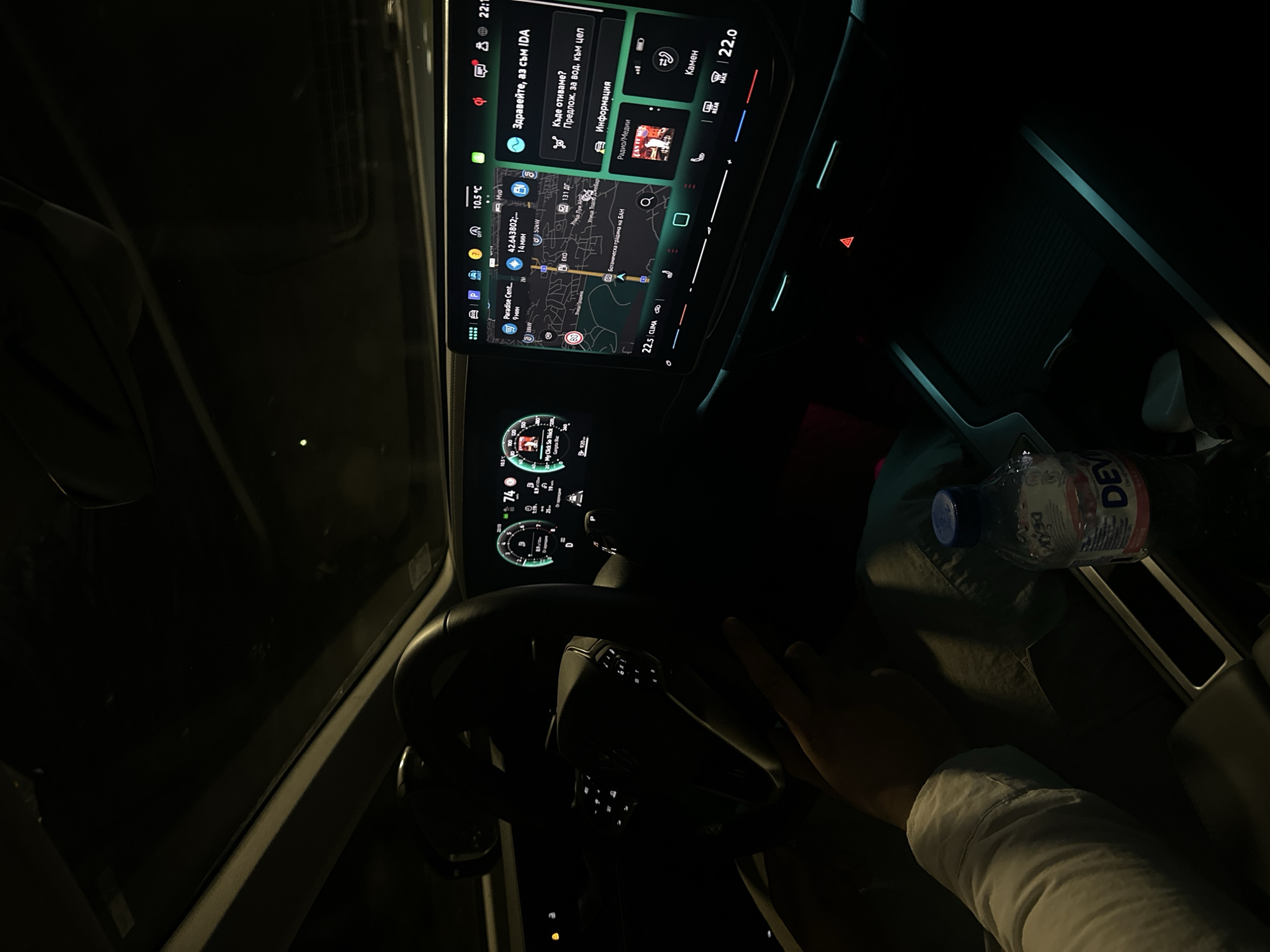 The image shows the interior of a car at night. The driver is visible, with their hand on the steering wheel and wearing a white shirt. The car's dashboard is illuminated, displaying navigation information, music details (artist name "Ganpta Bac" and title "My Click So Thick"), and vehicle status. The navigation screen shows the car's location near "Ботаническа градина на БАН" (Botanical Garden of the Bulgarian Academy of Sciences) in Sofia, Bulgaria. A water bottle is in the passenger area. The outside temperature displayed on the dashboard is 10.5°C.