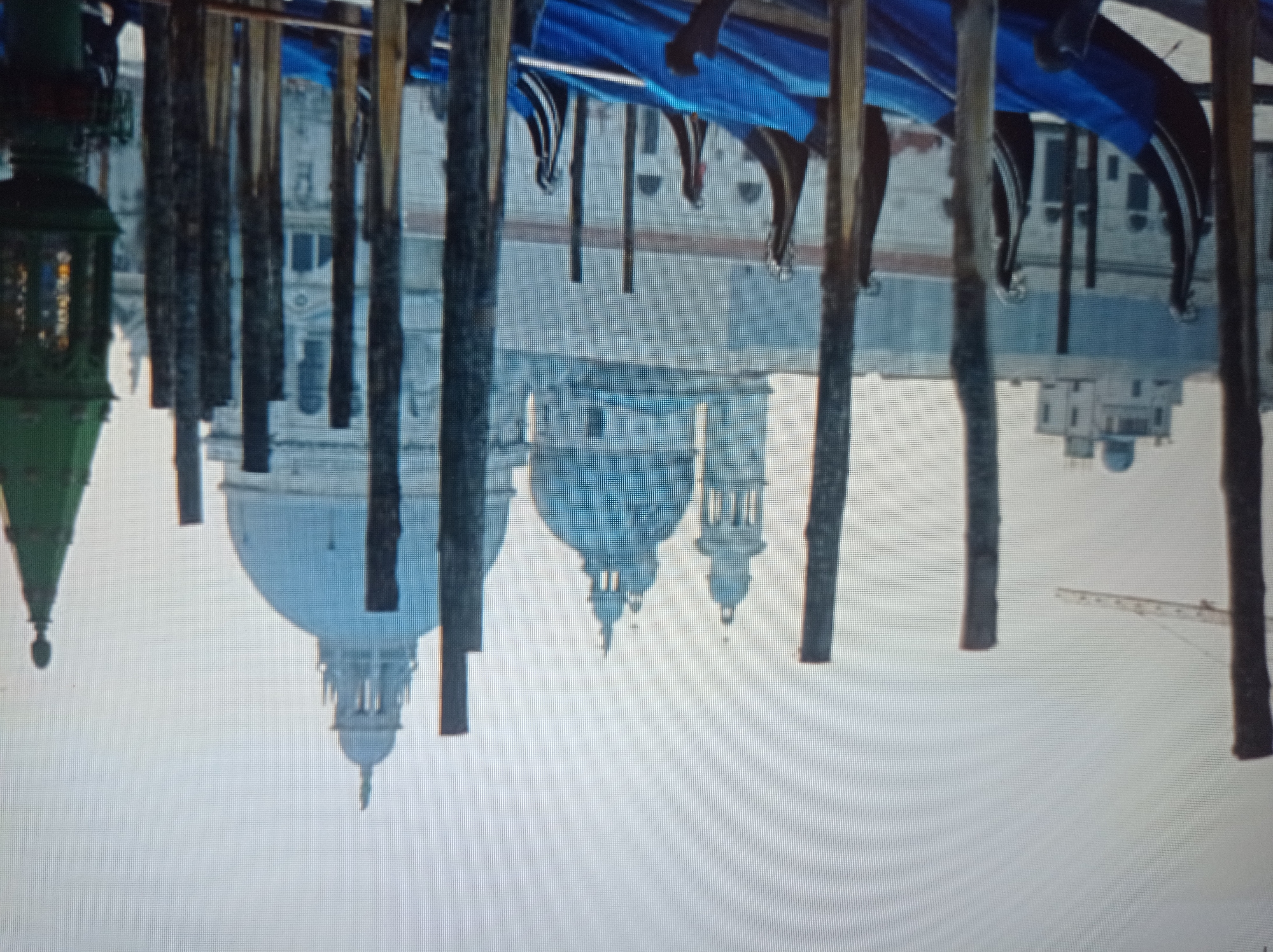 The image depicts a waterfront scene with several gondolas in the foreground, their prows adorned with decorative carvings. The gondolas are covered with blue tarps. Behind the gondolas, a collection of buildings with domes and spires are visible, suggesting a city with significant historical architecture. In the far background, a construction crane can be seen. The sky is overcast. This scene is not located in Mukachevo, Ukraine, but rather appears to be in Venice, Italy, known for its canals and gondolas.