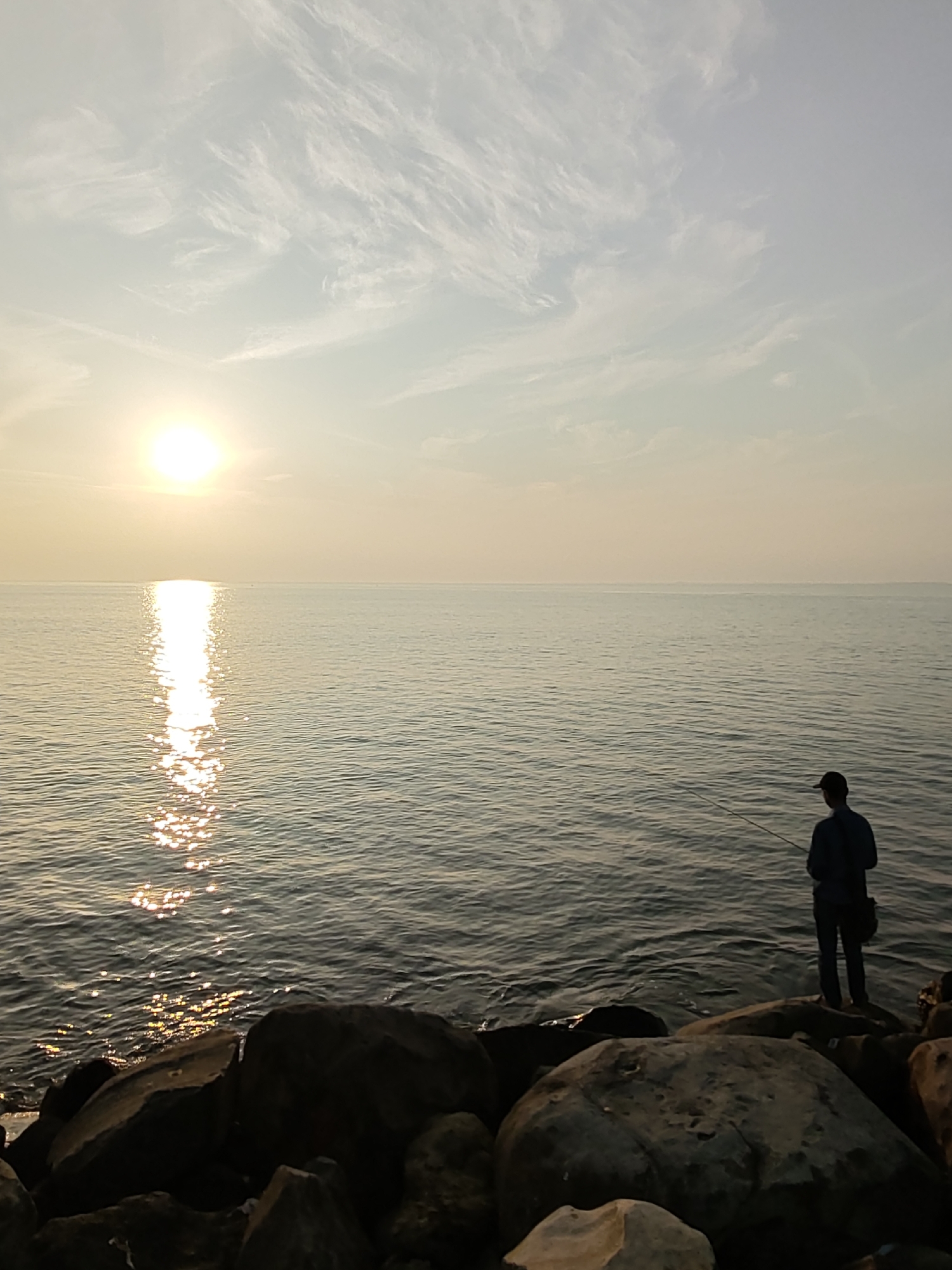 The image depicts a serene coastal scene at sunset, likely located in Lhokseumawe, Indonesia. The sun, a bright orb near the horizon, casts a shimmering reflection across the calm water. Wispy clouds streak across the pale sky, softening the transition between day and night. In the foreground, large rocks line the shore. A lone figure, silhouetted against the bright sky, stands on the rocks and appears to be fishing, holding a fishing rod. The person is wearing a dark long-sleeved shirt, pants, and a cap, with a bag slung over their shoulder. The overall atmosphere is peaceful and tranquil, with the warm hues of the sunset dominating the scene.