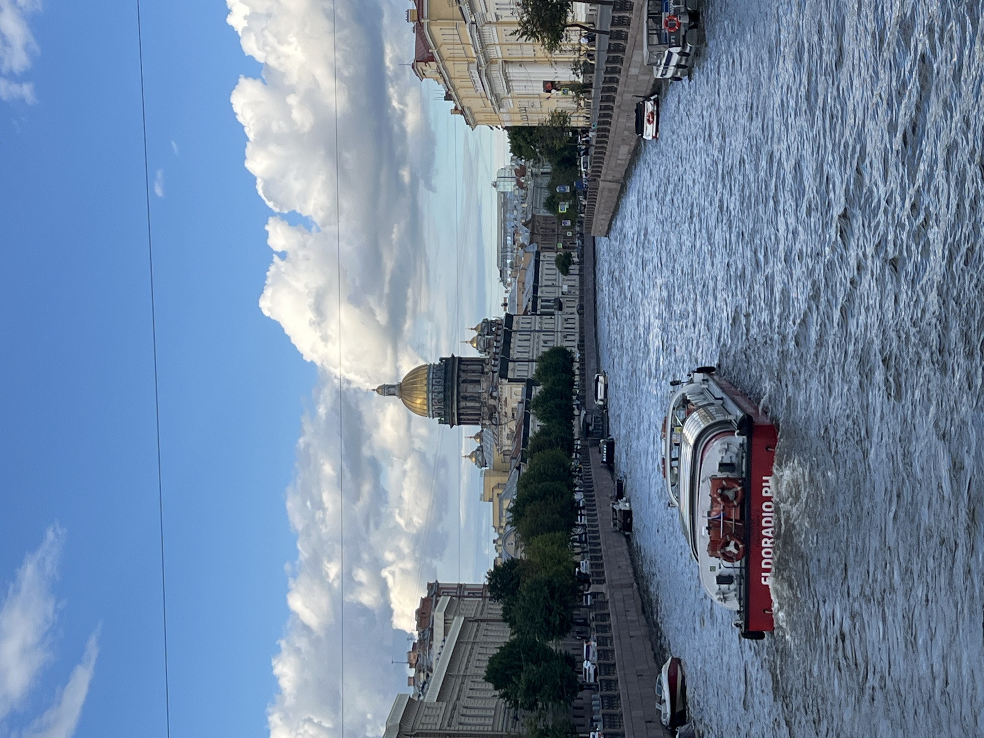 The image depicts a view of a wide canal in Saint Petersburg, Russia, with a large boat in the foreground and the iconic Saint Isaac's Cathedral in the background. The boat, which is red and white, has "ELDORADIO.RU" printed on its side. It is moving away from the viewer, creating ripples in the water. Along the canal banks are buildings with classical architecture, trees, and other smaller boats docked. The sky is partly cloudy, with dramatic white clouds against a blue backdrop. The overall scene suggests a leisurely day on the water, likely a tourist excursion.