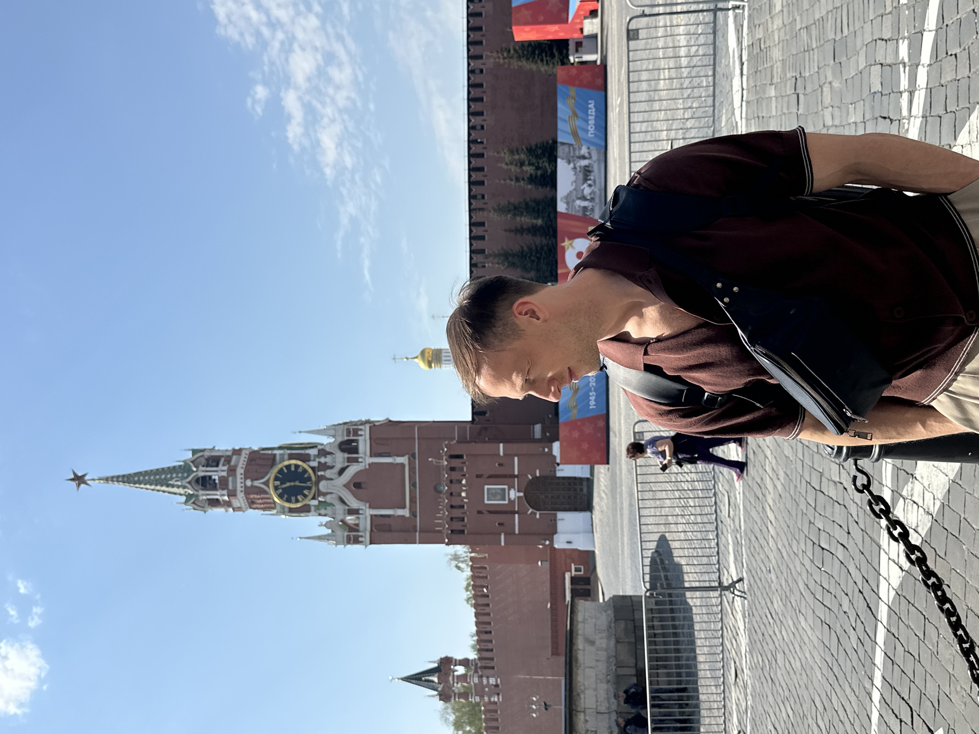 A man stands in Red Square, Moscow, facing the Kremlin, its distinctive spires, clock tower, and red brick walls visible in the background. He wears a brown polo shirt and a black shoulder bag. A metal fence and chain barrier are near him. Other people and banners are in the background. The sky is blue with scattered clouds. The ground is cobblestone pavement.