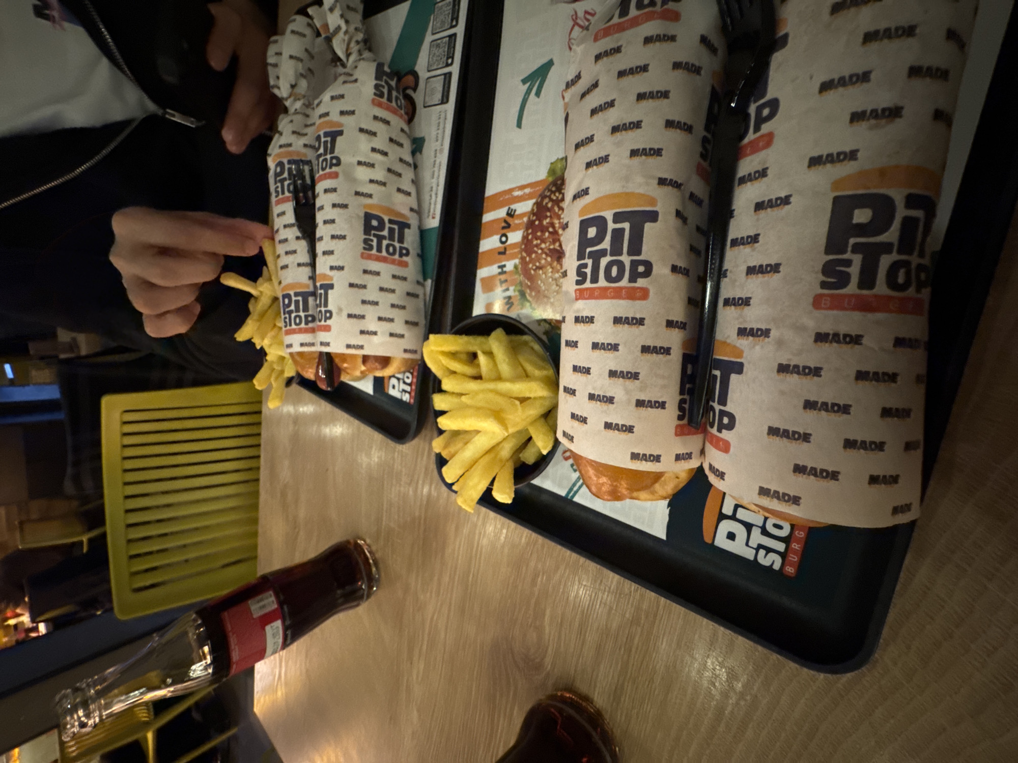 This image captures a close-up, overhead view of a fast-food meal at an indoor restaurant, likely in Yerevan, Armenia. The primary subjects are two trays laden with food, a person's hands, and beverages.
On a light wooden table, two black trays are visible. Each tray contains a meal from "Pit Stop Burger," featuring items wrapped in distinctive white paper. This wrapping prominently displays the "PIT STOP BURGER" logo in dark blue and orange, along with the word "MADE" repeated extensively across the surface. One tray holds a black bowl of golden-brown french fries, with a person's hand reaching in to grab a fry. Another bowl of fries is partially visible on the second tray.
A clear glass bottle, possibly of a dark-colored soft drink like Coca-Cola, lies horizontally on the table. Part of a person wearing a dark jacket is visible on the left side of the frame, indicating they are seated at the table. In the background, a bright yellow slatted object, possibly a chair or part of the decor, is present.
Notable text includes "PIT STOP BURGER" as the prominent branding. The word "MADE" is seen countless times on the wrapping paper, emphasizing the freshness of the food. "WITH LOVE" is also discernible on a paper insert on one of the trays. The lighting is artificial, common for indoor dining, and reflections are visible on the table and bottles. The scene portrays a casual dining experience, likely during an evening meal.