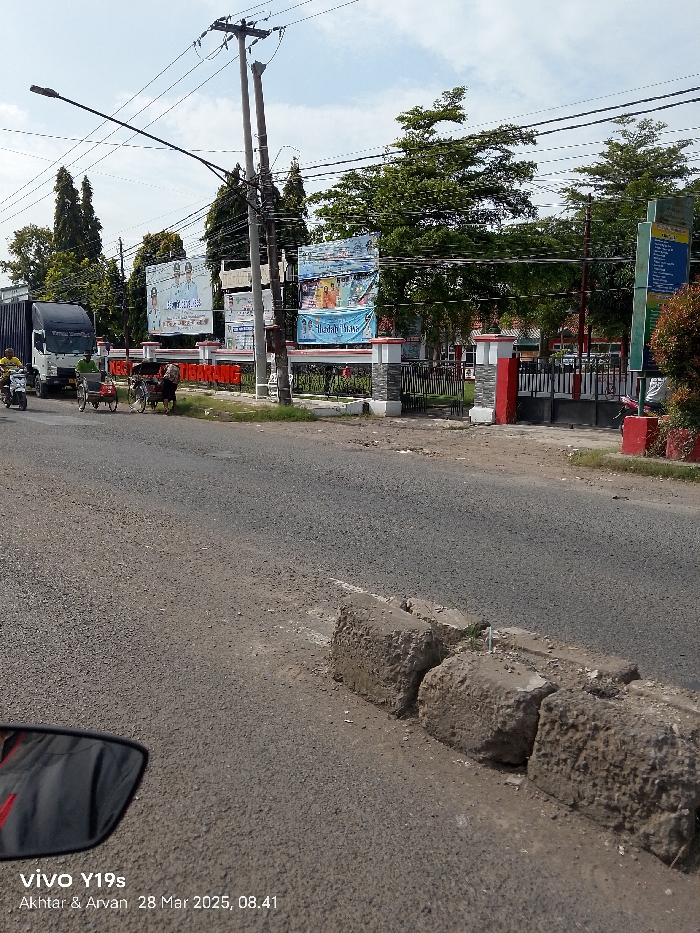 The image shows a street in Jatibarang, Indonesia, taken from a car traveling on the road. The road is paved with asphalt and has a white painted line down the center. There are four concrete blocks lining the right side of the road. The road is empty. The image is taken from a low angle, and the focus is on the road. There is a white truck parked on the left side of the road. In the background, there are several buildings with red-painted roofs and a large gate. The building in the center has a large billboard on its facade. The building on the right is partially obscured by lush greenery.