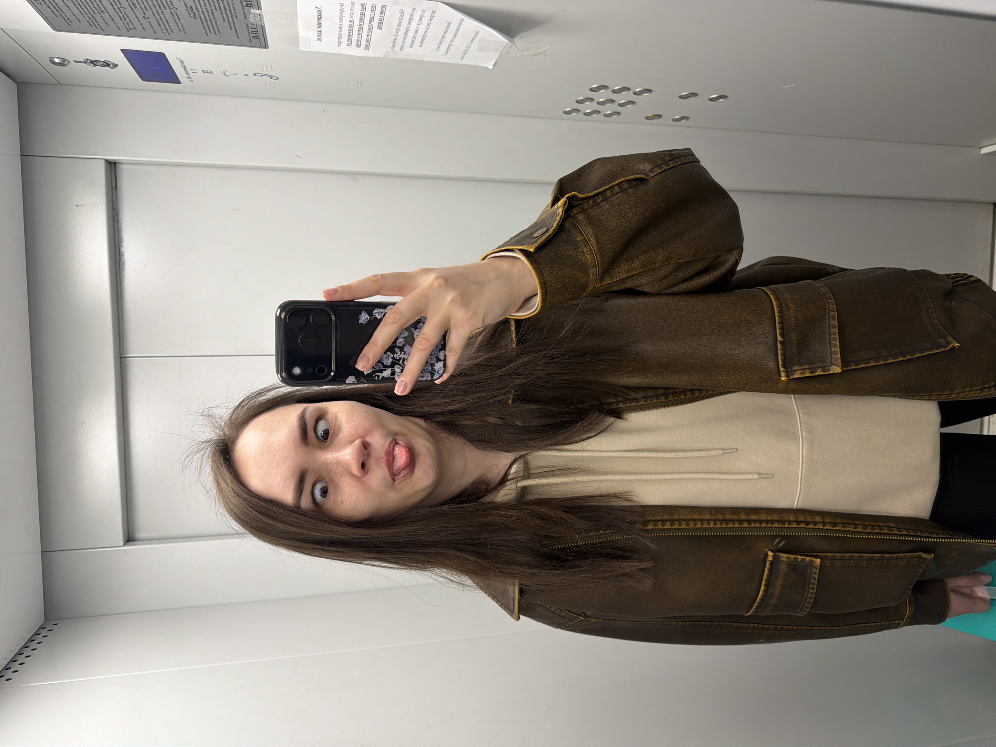 A young woman stands inside a light grey elevator taking a selfie. She is wearing a brown jacket over a beige hoodie and black pants. Her hair is dark brown and shoulder-length. She is making a face with her eyes wide and tongue sticking out. She holds a black cell phone in her right hand with a floral design on the case. The elevator walls are a plain light grey, with tactile Braille buttons and notices attached. No information about the city of Chaikovsky, Russia can be inferred from the image.