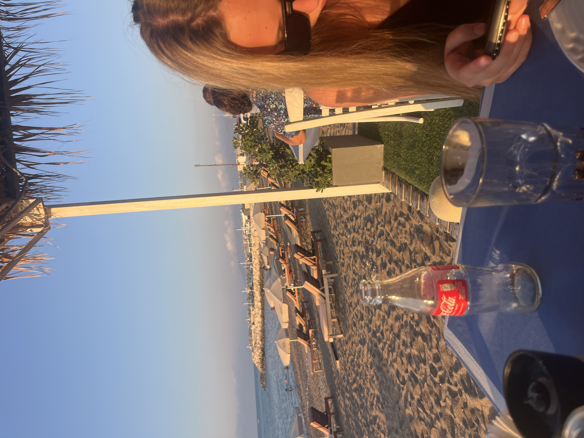 The image depicts a beach scene at sunset in Polis Chrysochous, Cyprus. In the foreground, a table with a blue surface displays a partially consumed bottle of Coca-Cola and a glass. A woman with long blonde hair, wearing sunglasses, is partially visible on the right side of the frame, holding a mobile phone. The background shows a sandy beach with numerous lounge chairs and umbrellas. The sea is calm with a few people swimming. A rocky breakwater extends into the sea on the left, and a coastline with buildings and vegetation is visible in the distance. The sky is clear and blue, indicating good weather. The overall atmosphere suggests a relaxed vacation setting.