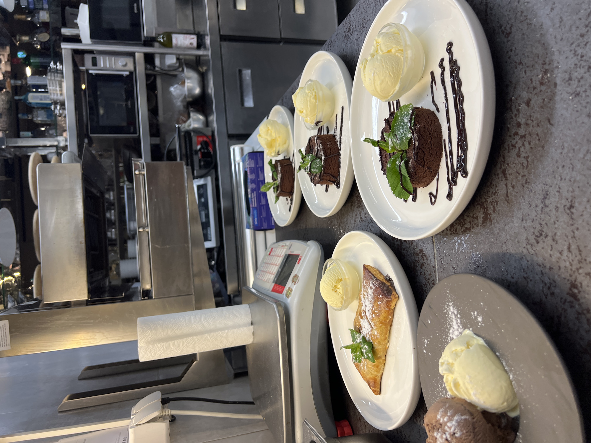 The image shows a kitchen counter with several plates of dessert. The desserts consist of chocolate cake, strudel, and ice cream, garnished with mint and chocolate sauce. The plates are arranged in a row on a dark countertop. In the background, there are stainless steel kitchen appliances such as ovens and refrigerators. A kitchen scale and paper towel dispenser are visible on the counter. The setting appears to be a commercial kitchen, likely in a restaurant or hotel. Location information is unavailable.