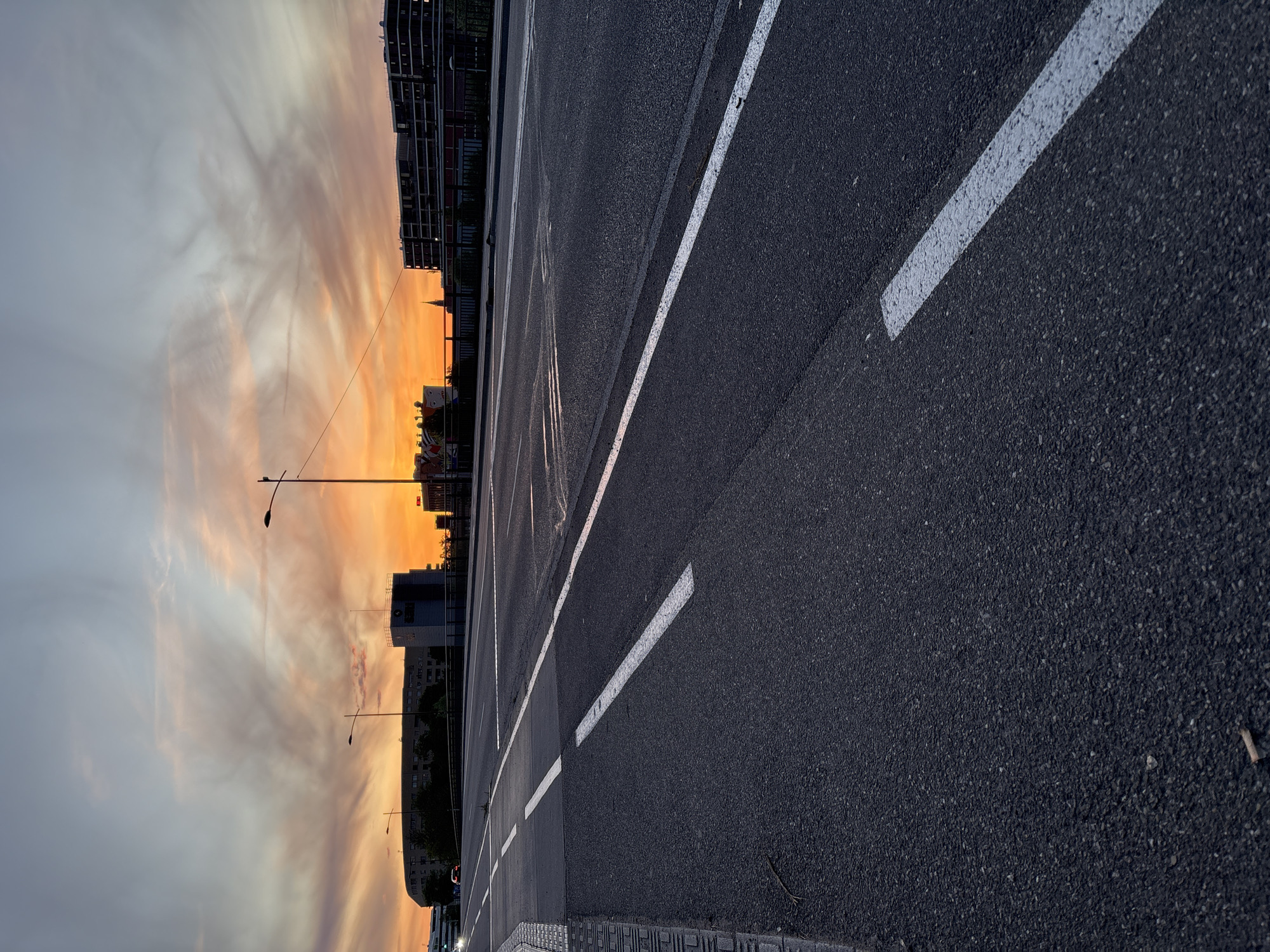 The image shows an asphalt road with white lane markings in Strasbourg, France. The road curves towards the background. On the left side of the image, there is a vibrant orange and gray sunset with buildings visible in the distance. Streetlights are also visible against the sunset. The focus is on the road surface and markings.