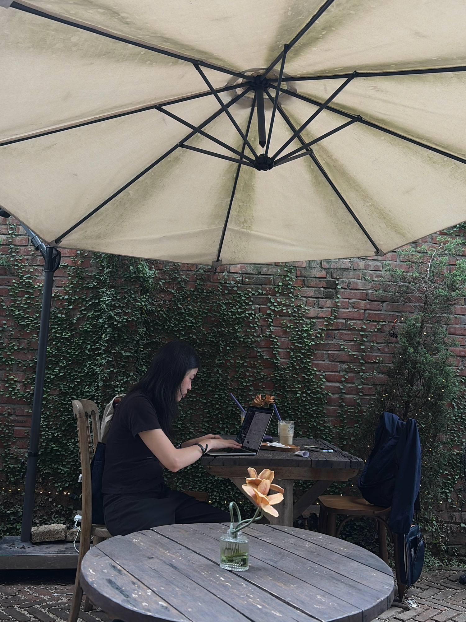 An outdoor cafe scene in Hanoi, Vietnam features a woman seated at a table, working on a laptop. She is wearing a black t-shirt and black pants. A beige umbrella is positioned overhead, providing shade. The table has a laptop, a glass of beverage, and a small vase with a flower. Behind the table, a brick wall is covered in lush green ivy. To the right, a small evergreen tree is decorated with subtle lights. A dark blue jacket is draped over the back of a chair. In the foreground, a round wooden table is visible. The ground is paved with small stones and brickwork.