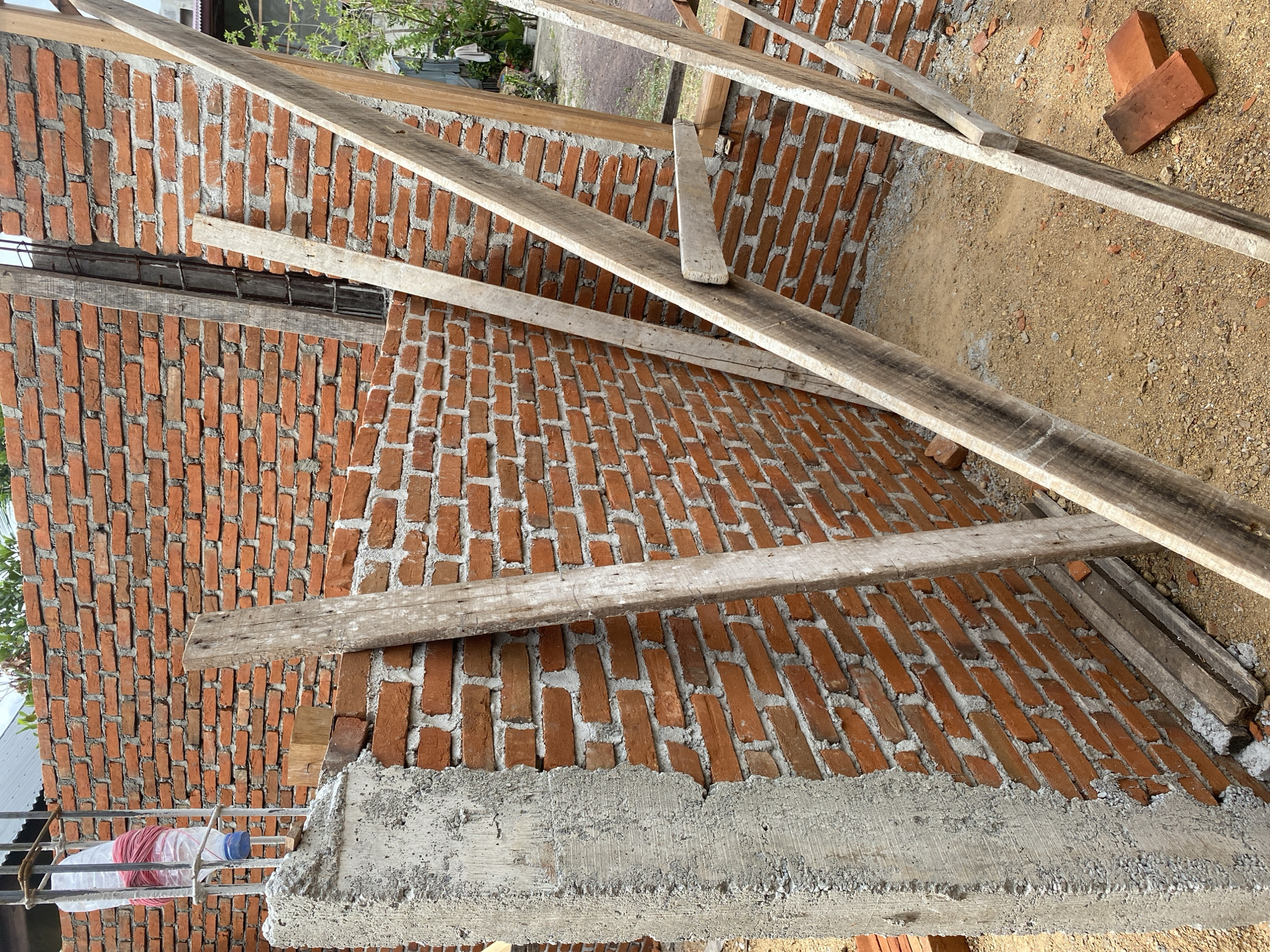 An outdoor, eye-level, medium shot captures a construction site focused on a brick wall under development. The wall, made of orange bricks and white mortar, occupies the majority of the frame. Several rough, unfinished wooden planks are propped against the wall, some diagonally, suggesting they are part of temporary scaffolding or bracing. In the foreground, a concrete pillar with visible rebar and a plastic bottle filled with red twine stands to the left. The ground is covered in sand and small stones, with a few loose bricks scattered around. The background reveals more of the brick wall, a glimpse of vegetation, and a corrugated metal roof, indicating an undeveloped or semi-developed area. The scene appears to be in an urban or suburban environment.