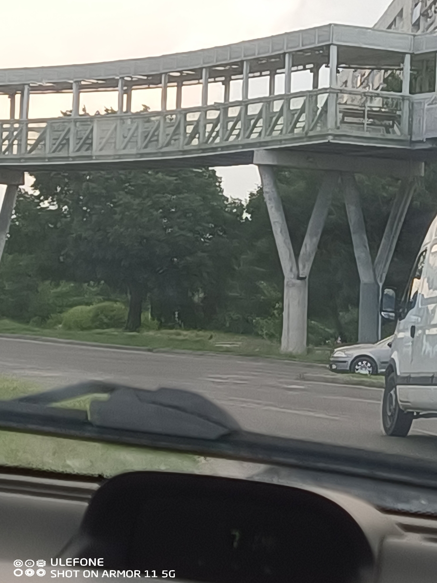 The image depicts an elevated pedestrian bridge over a road in Burgas, Bulgaria, specifically the bridge spanning Transportna Street. The bridge, constructed from light-colored concrete or metal, features a covered walkway with an open side structure resembling a series of windows, and distinctive 'A-frame' or zigzag railings. It is supported by large, Y-shaped concrete pillars. Multiple human figures are discernible as dark silhouettes walking along the bridge. Below, a paved road extends across the midground. A white van is partially visible in the foreground on the right, and a silver sedan is parked or stopped further down the road on the left. Dense green foliage and several trees line the road and extend into the background. The sky above is bright and hazy. The photograph was taken from inside a vehicle, with the dashboard, windshield wipers, and a section of a dark car seat visible in the foreground. A digital display on the dashboard shows the number '7'.