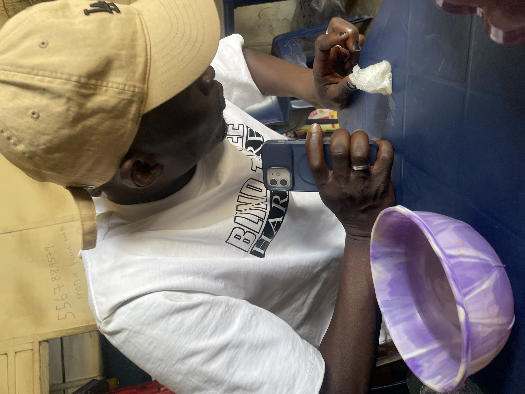 An overhead shot shows a dark-skinned individual wearing a tan baseball cap with a black logo, a white T-shirt with black lettering, and a ring on their left ring finger. They are using a blue smartphone to take a photo of a small piece of white material resting on a blue surface. A purple and white plastic bowl is next to the smartphone. In the background, a yellow wall has "5567889579. Mobile Point Jos." written on it. The scene is located in Jos, Nigeria.