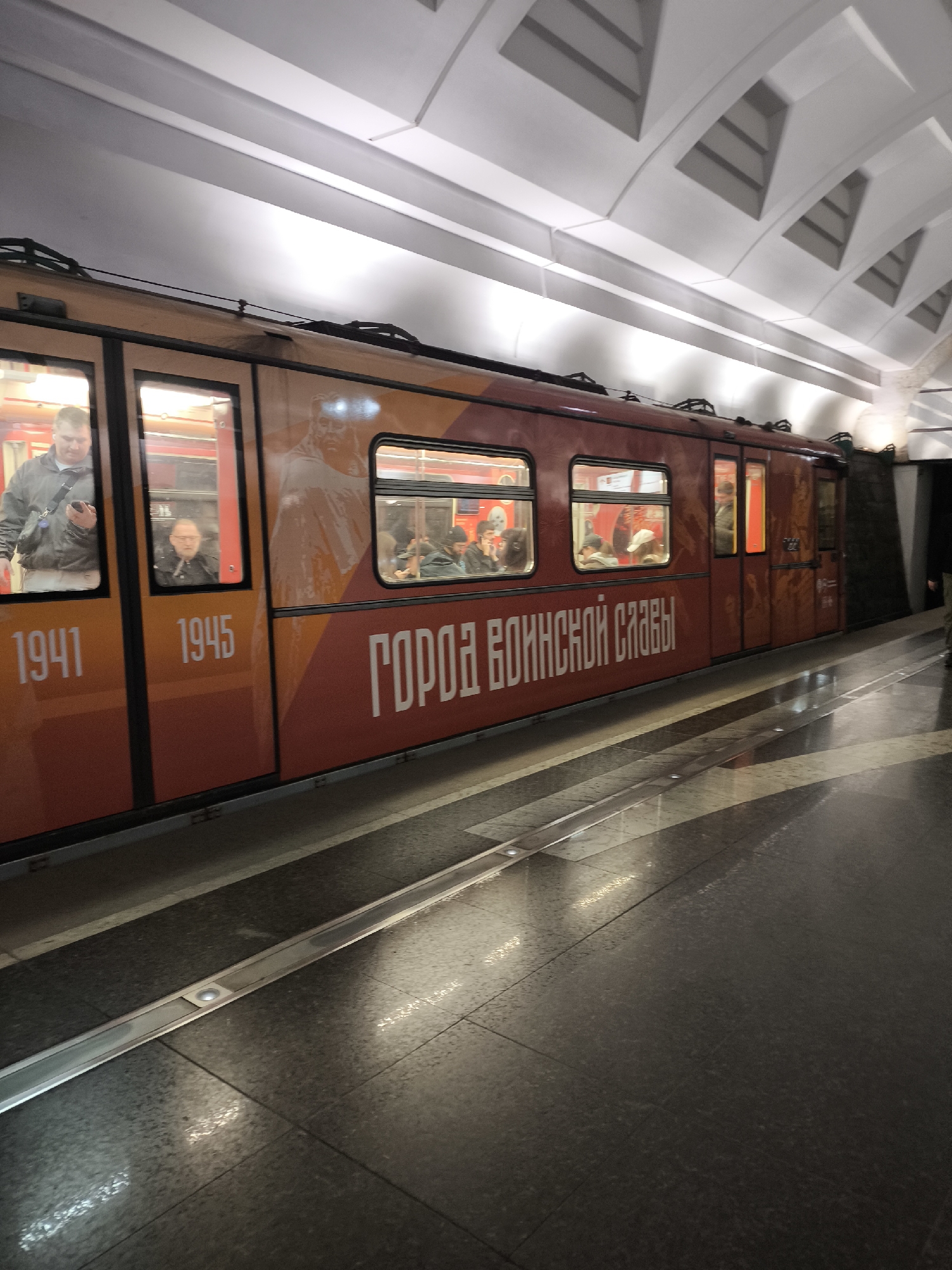 The image shows a train at a subway station in Unknown, Unknown. The train cars are decorated with the years 1941 and 1945, a portrait, and the inscription "Город воинской славы". People are visible inside the train. The subway station has a glossy tile floor and a modern white ceiling.