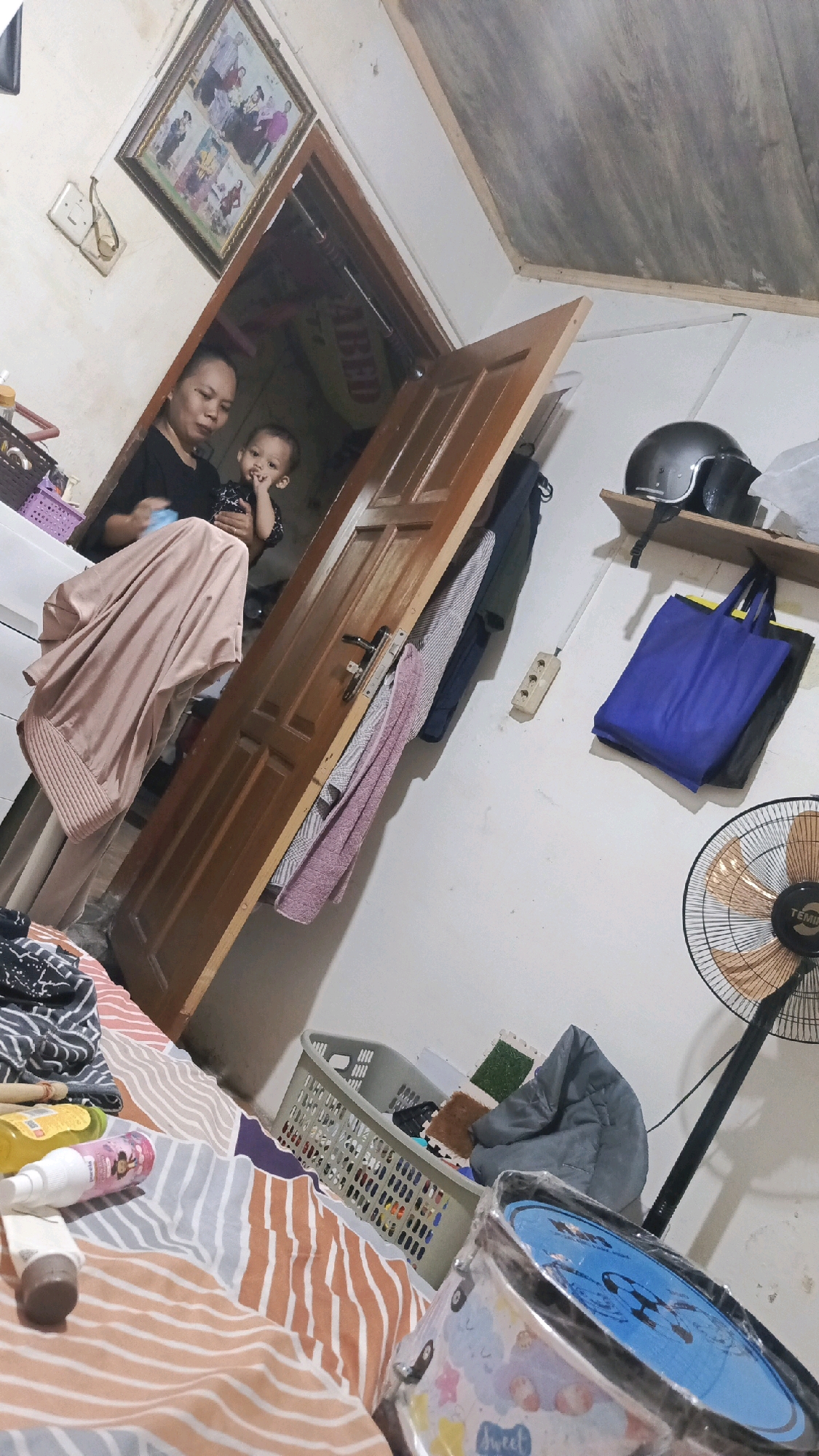 The image depicts an indoor scene, likely a bedroom, in a modest dwelling. The perspective is tilted, suggesting the photo was taken at an unusual angle.
In the foreground, a bed with a striped orange and white comforter is visible. Several items are placed on the bed, including toiletries like a white spray bottle and a brown-capped tube, and a yellow bottle. To the right, partially in frame, is a cylindrical object, possibly a toy drum, wrapped in clear plastic.
Further back, a light gray laundry basket filled with items is positioned on the floor. Beside it, a folded gray blanket or cushion rests.
A dark wooden door stands open, revealing a glimpse into another room. On the door itself, several articles of clothing are draped, including a pink and white striped towel or blanket and a dark blue garment.
To the left of the door, a woman in a black shirt and light-colored pants is holding a young child. The child is looking towards the camera. Above them, a framed picture hangs on the wall, displaying multiple family portraits.
On the right side of the room, a wooden shelf is mounted on the wall. On the shelf, a dark gray helmet and a black bag are placed. Below the shelf, a blue tote bag is hanging. A black standing fan with a circular grill is positioned on the floor to the right.
The walls appear to be painted white, and there are light switches and electrical outlets visible. A wooden-slatted ceiling is also partially visible. The overall impression is of a lived-in space with everyday objects. The reference to Pekauman Kulon, Indonesia, suggests a specific location within a residential area.