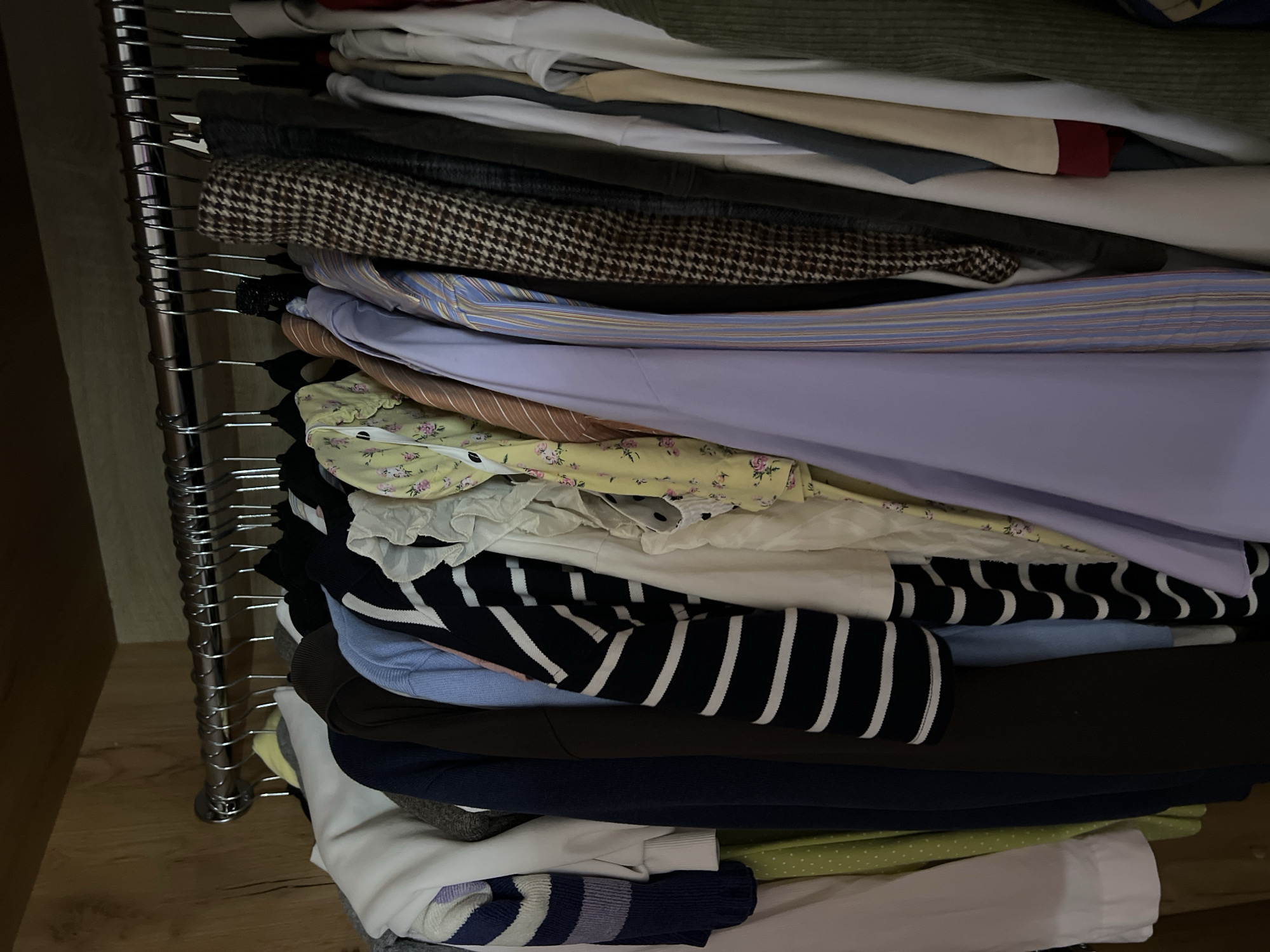 A closet filled with clothing hanging on a metal rod. The clothing consists of various garments including sweaters, shirts, and dresses in different colors and patterns such as stripes, floral prints, and houndstooth. The closet appears to be made of wood.
