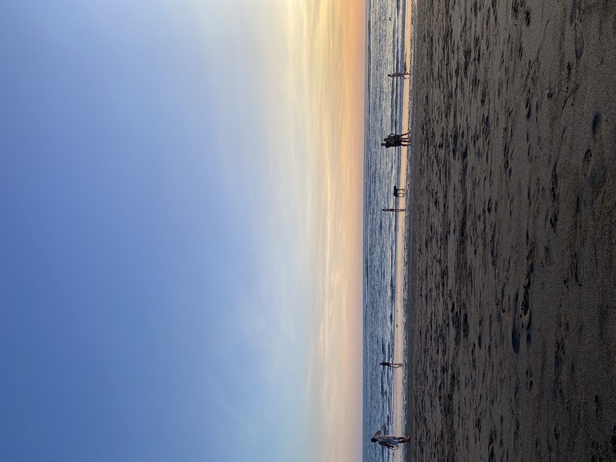The image shows a beach scene in North Kuta, Indonesia, likely during sunset. The sky transitions from blue to orange near the horizon. The ocean is visible in the middle, with gentle waves. Several people are seen along the shoreline, some in groups and others alone. A dog is present near one group of people. The beach sand is dark, with numerous footprints.