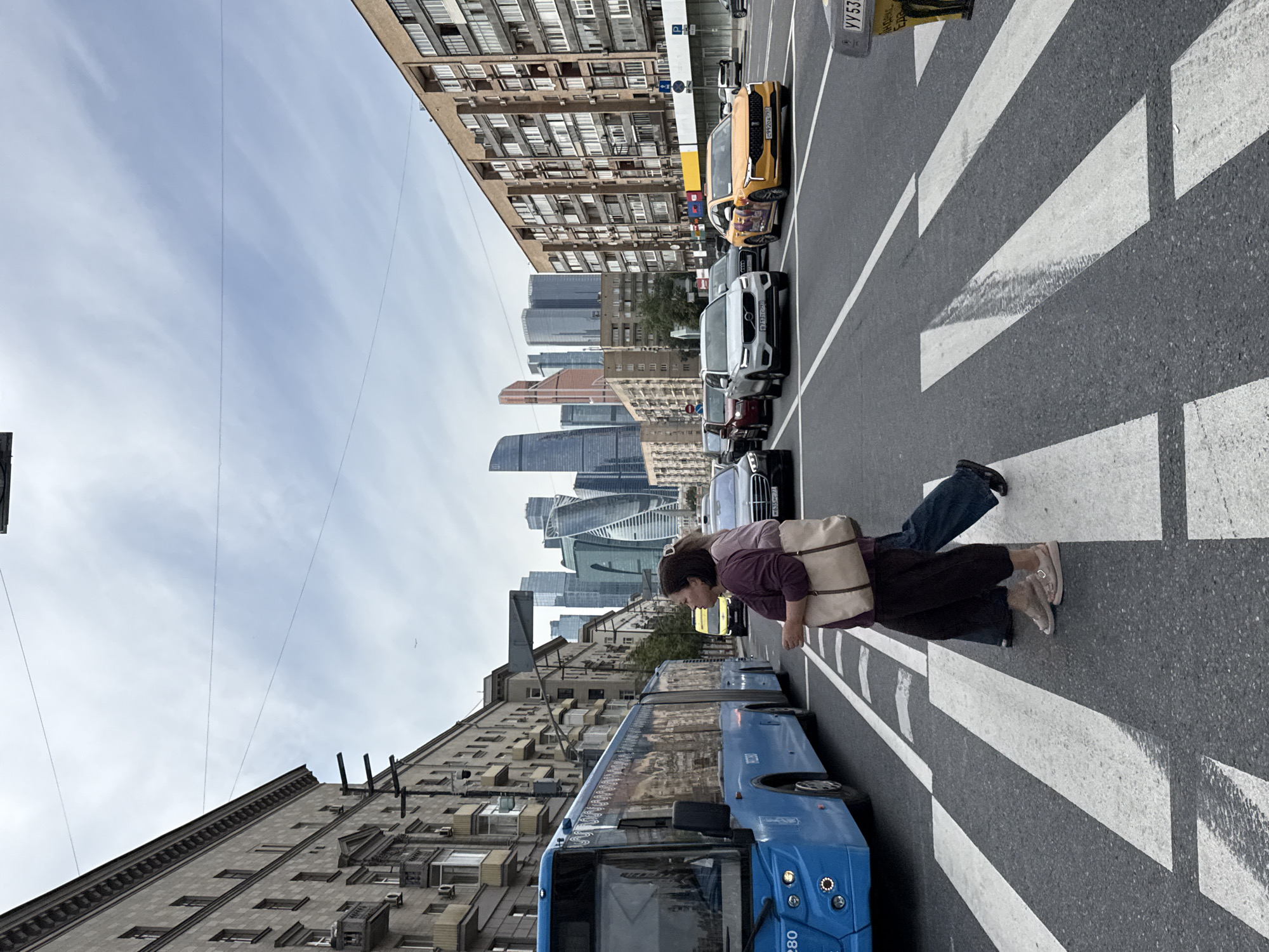 This is a street-level, eye-level, full-frame shot capturing a crosswalk scene in Moscow, Russia, with the Moscow City business district skyscrapers visible in the background. In the foreground, a woman in a purple top and dark pants, carrying a beige tote bag, is crossing the street from right to left. To her left and slightly behind, another person in blue jeans and dark shoes is also crossing. A blue articulated bus is stopped to the left, partially blocking the view of the street. Several cars are also present on the road, including a yellow taxi on the right and a white SUV. The buildings on either side of the street are multi-story residential and commercial structures. The sky is overcast with scattered clouds.