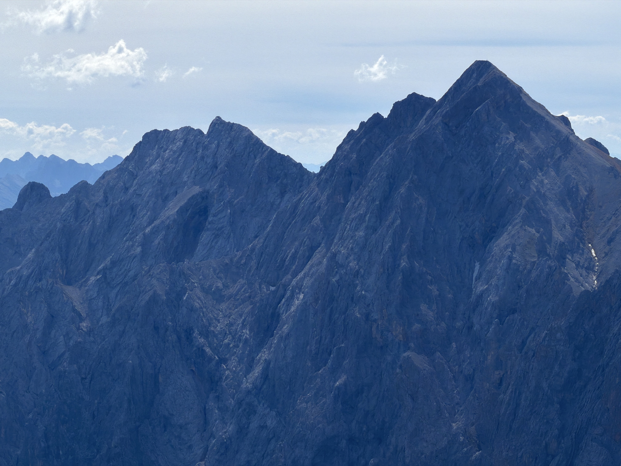 The image depicts a vast mountain range under a hazy sky with scattered clouds. The mountains are predominantly dark blue, indicating a rugged, rocky terrain with steep slopes and sharp peaks. The lighting suggests it is daytime, but the atmosphere is somewhat muted, creating a sense of distance and depth. In the foreground, jagged rock formations dominate the view, with visible textures of scree and weathered stone. Further in the distance, the mountain peaks become softer and bluer, suggesting they are farther away. The sky is a pale blue with patches of white clouds, some of which are wispy and others more defined. There are no visible signs of human presence, structures, or any specific landmarks that would pinpoint a precise location within Garmisch-Partenkirchen without further context or information. The overall scene is one of natural grandeur and rugged alpine scenery.