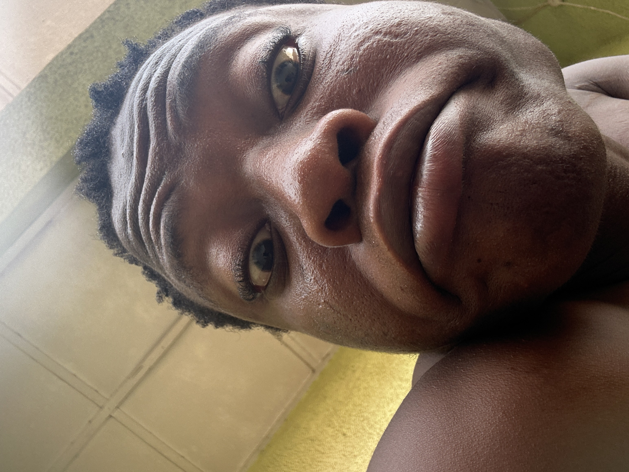 A close-up, low-angle shot depicts a dark-skinned individual's face and upper body. The person's head is tilted back, and their facial expression appears somewhat contorted, with raised eyebrows and a pursed mouth. The person is indoors, with a yellow painted wall visible in the background. The image location is unknown.