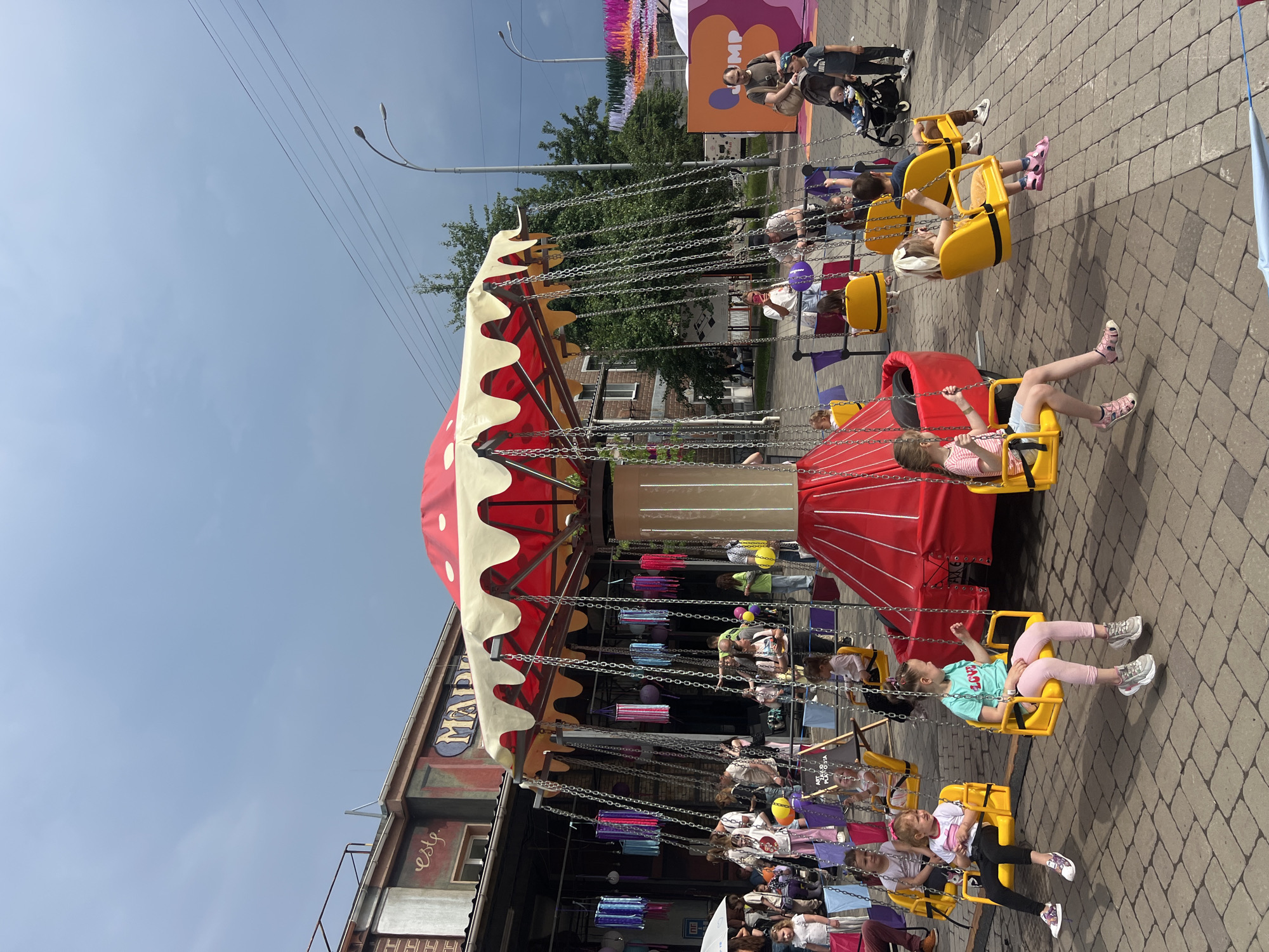 This image depicts an outdoor daytime scene at what appears to be a fairground or event space, specifically identified as Art Zavod Platforma in Kyiv, Ukraine. The central focus is a stationary swing carousel with a distinctive red and white mushroom-shaped canopy with yellow scalloped trim, supported by a central light-colored column and a red base. Multiple yellow single-seat swings, attached by metal chains, are positioned around the carousel.
Approximately ten children, predominantly girls, are seated in the yellow swings, some with their legs dangling, others resting their feet on the ground or the swing structure. They are dressed in casual summer clothing including t-shirts, shorts, and sandals. Several adults are present in the scene; some are standing near the carousel, observing the children, while others are walking in the background. One adult on the right is pushing a dark-colored baby stroller containing an infant, while another young child stands nearby. Another adult is visible in the middle-left, holding a white umbrella.
The ground is paved with light brown interlocking brick pavers. In the background, on the left, a multi-story building with a rustic, brick-and-stucco facade is visible, featuring faded signage that includes the word "este" and a partial "MAP" (likely part of "Art Zavod Platforma"). Colorful fabric streamers and lanterns in various shades of blue, pink, purple, and yellow hang as decorations from structures near the building. Further in the background on the right, additional colorful streamers are visible. Green trees with dense foliage are interspersed throughout the background. A street light with two luminaires and overhead power lines extends into the frame on the right side of the image. The sky is overcast with a uniform light grey-blue tone. A large, colorful sign with the word "JUMP" is partially visible in the far right background.