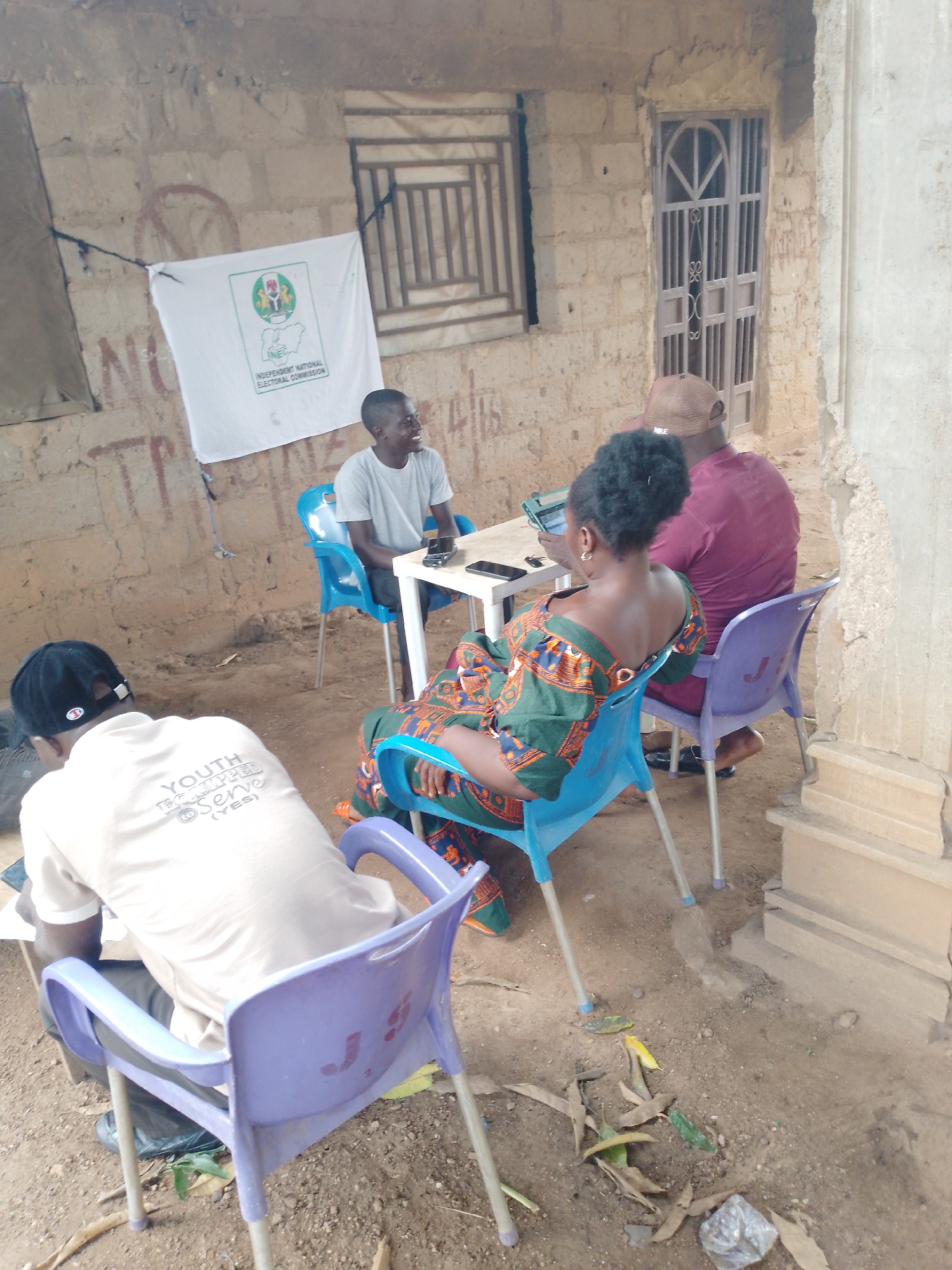The image depicts an outdoor scene, possibly a makeshift polling station or meeting area, in an urban environment. Four individuals are present. In the foreground, a person wearing a white t-shirt with the inscription "YOUTH EQUIPPED SERVE (YES)" and a black baseball cap is seated, facing away from the camera, with papers in front of them. To their right, a woman wearing a colorful patterned dress is seated on a blue chair, looking towards a table where a man in a maroon shirt and a tan cap is seated. The man is looking at a tablet. A white banner with the logo of the Independent National Electoral Commission (INEC) is displayed on the wall behind the man in the maroon shirt. The environment consists of a dirt ground, a rough stone wall, and a barred doorway in the background. Fallen leaves are scattered on the ground.