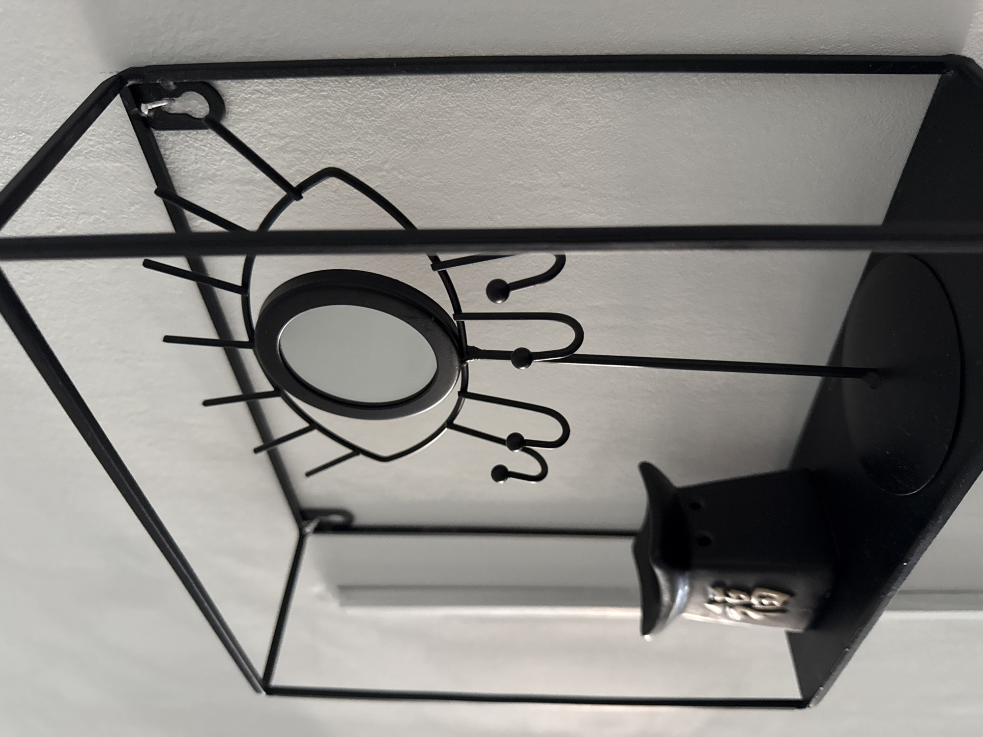 An upward-angled shot captures a black, decorative metal object against a textured, light-colored ceiling. The object features a geometric, possibly hexagonal, outer frame made of thin metal rods. Within this frame, a central circular element, resembling a small mirror or a light fixture, is surrounded by stylized metal extensions that curl and end in small spherical tips, resembling hooks or rays.
The object is mounted to the ceiling or upper wall, as indicated by a silver screw or nail visible through a keyhole-shaped mounting slot in the top left corner of the frame. The background is a uniformly lit, off-white or light gray surface with a fine, granular texture.
In the lower right portion of the image, another black, structured object is partially visible, appearing to be a separate decorative piece or an integral part of the main display. This secondary object has a flat base and a more solid, sculpted shape. Embossed characters, possibly "1G6" or similar, are visible on its surface, but are somewhat obscured and challenging to read clearly. The overall scene depicts an indoor decorative arrangement.