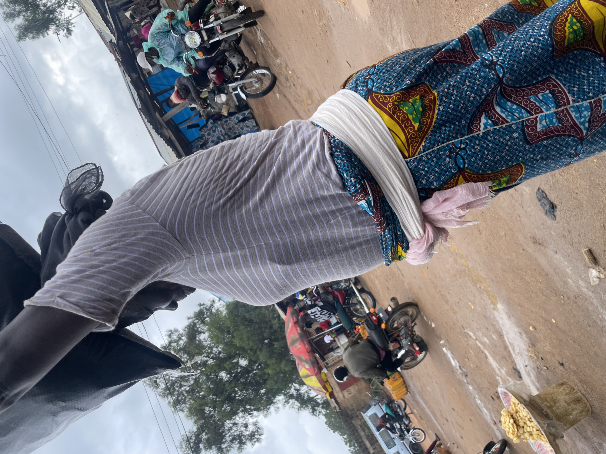A medium shot captures a person from the chest down, carrying a large, dark bag on their head. The individual wears a horizontally striped long-sleeved shirt and a vibrant, patterned skirt with a white sash tied around their waist, with a pink scarf peeking out. The scene unfolds on a dirt road in what appears to be a bustling market in Wang, Nigeria. Several motorcycles are visible, with some people riding them and others standing around. In the background, market stalls with corrugated metal roofs line the street, suggesting a commercial area. The ground is wet and muddy, with puddles of water indicating recent rain. The sky is overcast, contributing to the somewhat dim lighting. A plate of food, possibly snacks or grains, is displayed on a makeshift stand in the foreground.
