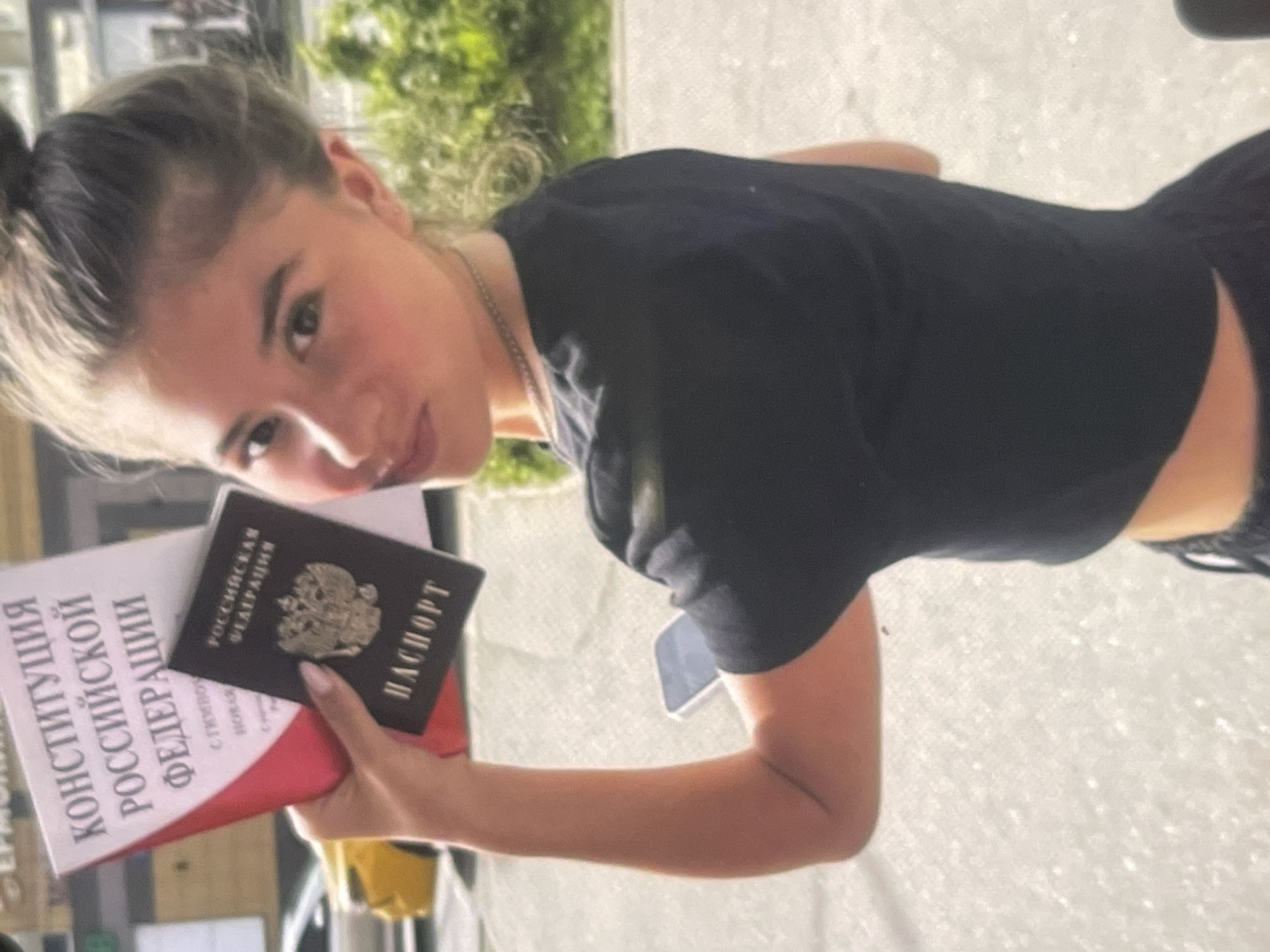 The image, rotated at an angle, features a young woman looking directly at the camera with a slight smirk. She has light brown hair, partially tied up, and is wearing a black short-sleeved crop top with a silver-colored chain around her neck.
In her left hand, she holds two official-looking documents. The top one is a dark-colored passport-like item, clearly displaying the emblem of the Russian Federation, the words "РОССИЙСКАЯ ФЕДЕРАЦИЯ" (Russian Federation), and "ПАСПОРТ" (Passport) in Cyrillic text. Beneath it, she holds a larger book, white with red accents, on which the title "КОНСТИТУЦИЯ РОССИЙСКОЙ ФЕДЕРАЦИИ" (Constitution of the Russian Federation) is prominently visible. Her fingernails are manicured with a light polish.
The background includes green foliage at the top, suggesting an outdoor or brightly lit environment. A light-colored, textured surface is visible behind her, with a yellow object (possibly a cup) and a dark mobile phone resting on it. The scene is well-lit, indicating it is daytime. The provided location context for this media file is Poltava, Ukraine, while the documents shown are Russian.