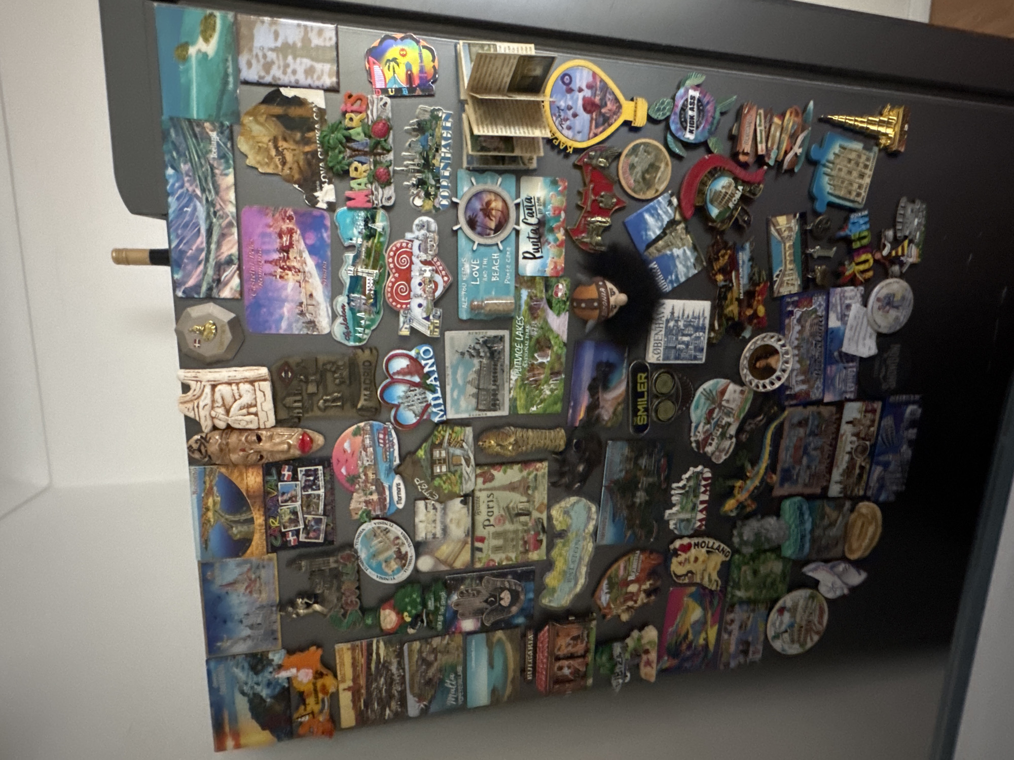 The image shows a collection of magnets densely covering a refrigerator door. The magnets appear to be souvenirs from various locations around the world, based on the names and images visible. Some of the locations mentioned include Paris, Tunisia, Marmaris, Madrid, Milan, France, Hungary (Balaton), Holland, Bulgaria, Malta, Copenhagen, and Rome. There is no indication in the image that any location is in the city of London, United Kingdom. The scene is indoors.