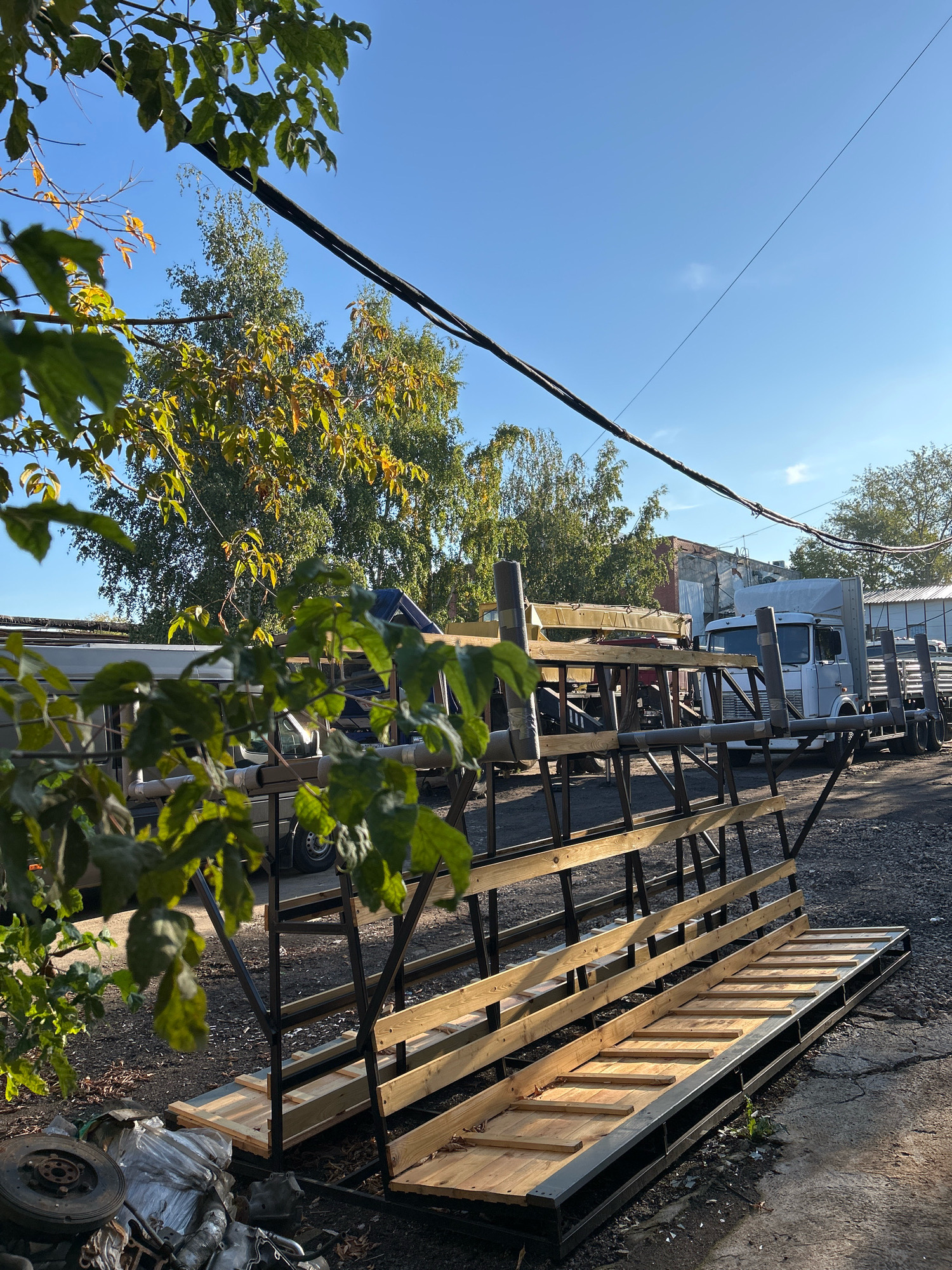 The image depicts an outdoor scene, likely a yard or a lot, with a clear blue sky overhead. In the foreground, a metal and wood rack structure is prominent. This structure appears to be unfinished, with wooden planks laid out on metal supports. To the left of the rack, a pile of discarded items, including what looks like a car tire and various metal parts, is visible.
Further back in the scene, several vehicles are parked. A white truck with a silver cargo box is the most prominent. Behind it, a yellow crane and other industrial vehicles or equipment can be seen. In the background, buildings with brick and metal siding are present, along with trees displaying autumnal foliage. Power lines stretch across the upper part of the frame. The ground is a mix of gravel and cracked pavement. The lighting suggests it is daytime, with sunlight casting shadows. No people are clearly visible in the frame.
Given the presence of industrial vehicles, storage racks, and a general utilitarian setting, this location could be a construction site, a vehicle repair yard, or a storage facility. Without further context, it's impossible to pinpoint a specific location within Moscow, Russia. However, the overall impression is one of an active, working outdoor space, possibly related to construction or logistics, in an urban or semi-urban environment.