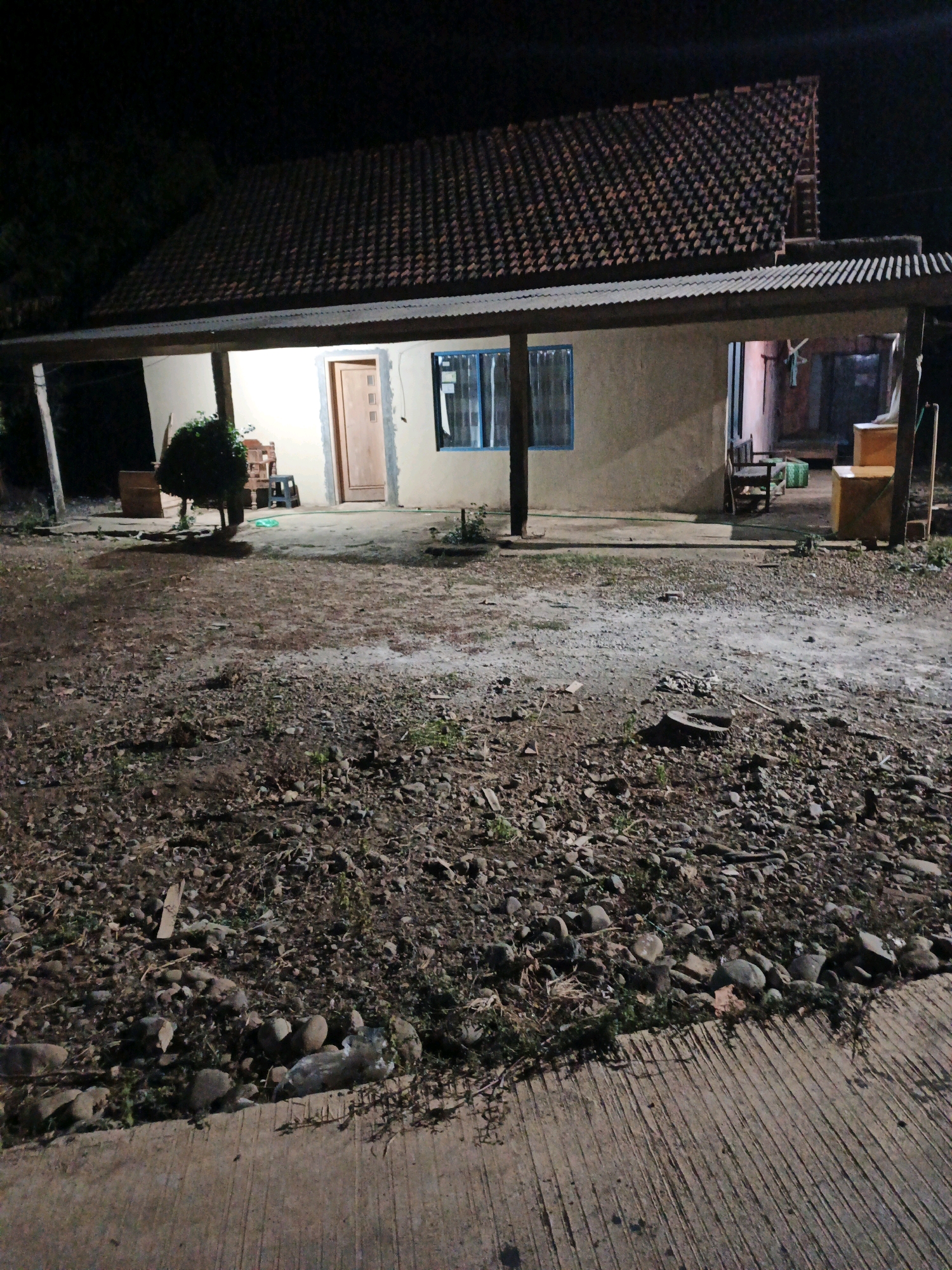 The image depicts the exterior of a house at night in Bumiharjo, Indonesia. The house has a tiled roof and a covered porch supported by wooden posts. A single door is visible on the front of the house, along with a window with blue trim. The ground in front of the house is unpaved and covered with dirt, stones, and debris. Some furniture, including chairs and boxes, is present on the porch and in the background. The scene is illuminated by artificial light, casting shadows. The environment appears to be rural or semi-rural. No people are visible in the image.