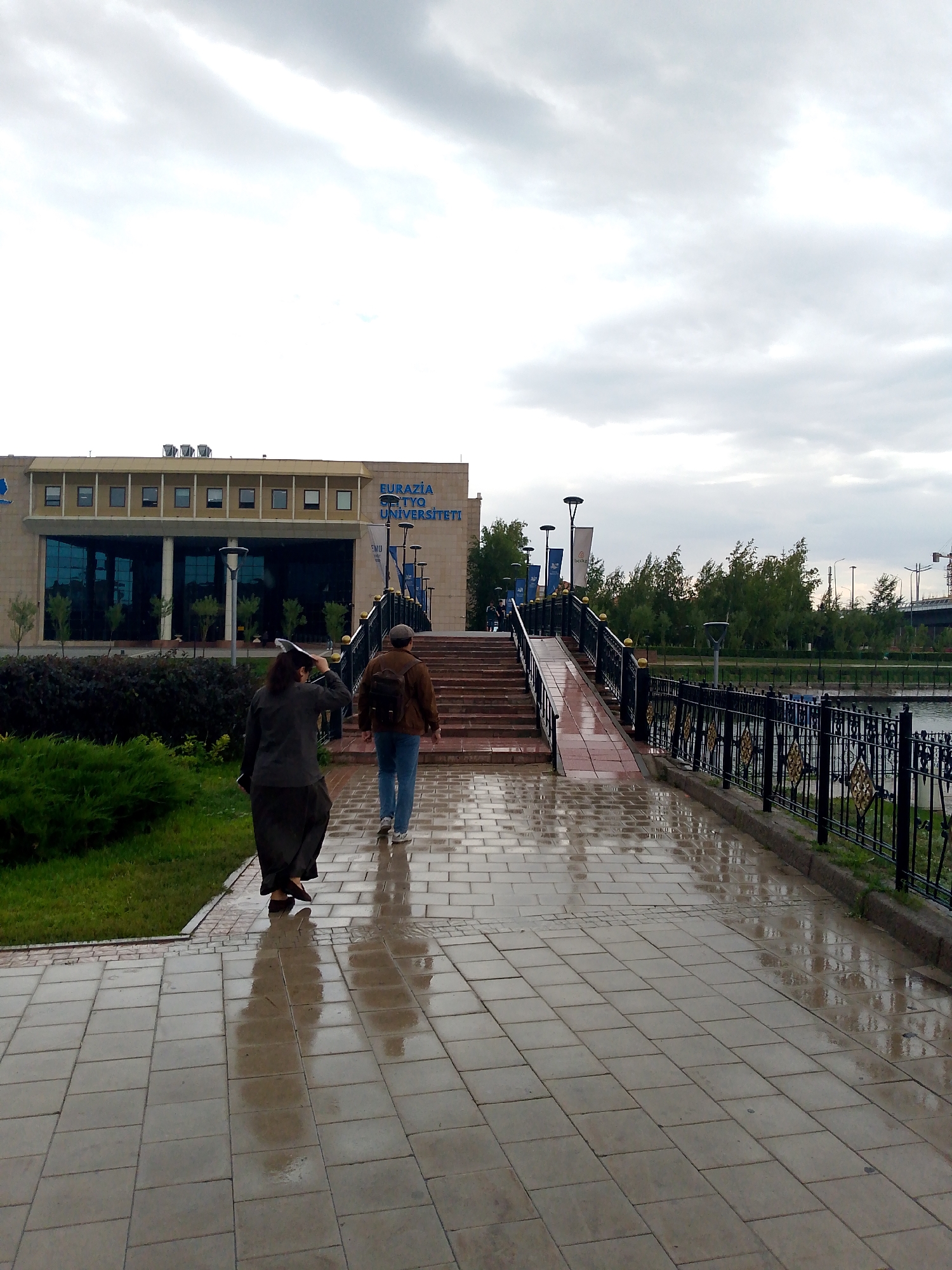 The image captures a rainy day scene in Astana, Kazakhstan, specifically in front of the Eurasian National University. Two individuals are walking away from the camera on a wet, tiled pathway. The person in the foreground, on the left, is wearing a dark long coat and skirt, holding a white object over their head. The person behind them, on the right, is a male wearing a brown jacket, blue jeans, and a backpack, walking up a ramp that leads to a set of stairs.
The architecture of the Eurasian National University is visible in the background. The building is modern, with a light-colored facade and large glass windows. Blue lettering on the building reads "EURASIA UNIVERSITY". The pathway and stairs are wet, reflecting the overcast sky and surrounding greenery. A black metal fence with gold embellishments lines the right side of the pathway, separating it from a body of water or parkland. Trees with green foliage are visible beyond the fence, and in the distance, a bridge and construction cranes can be seen. The overall atmosphere is subdued due to the cloudy and wet weather.