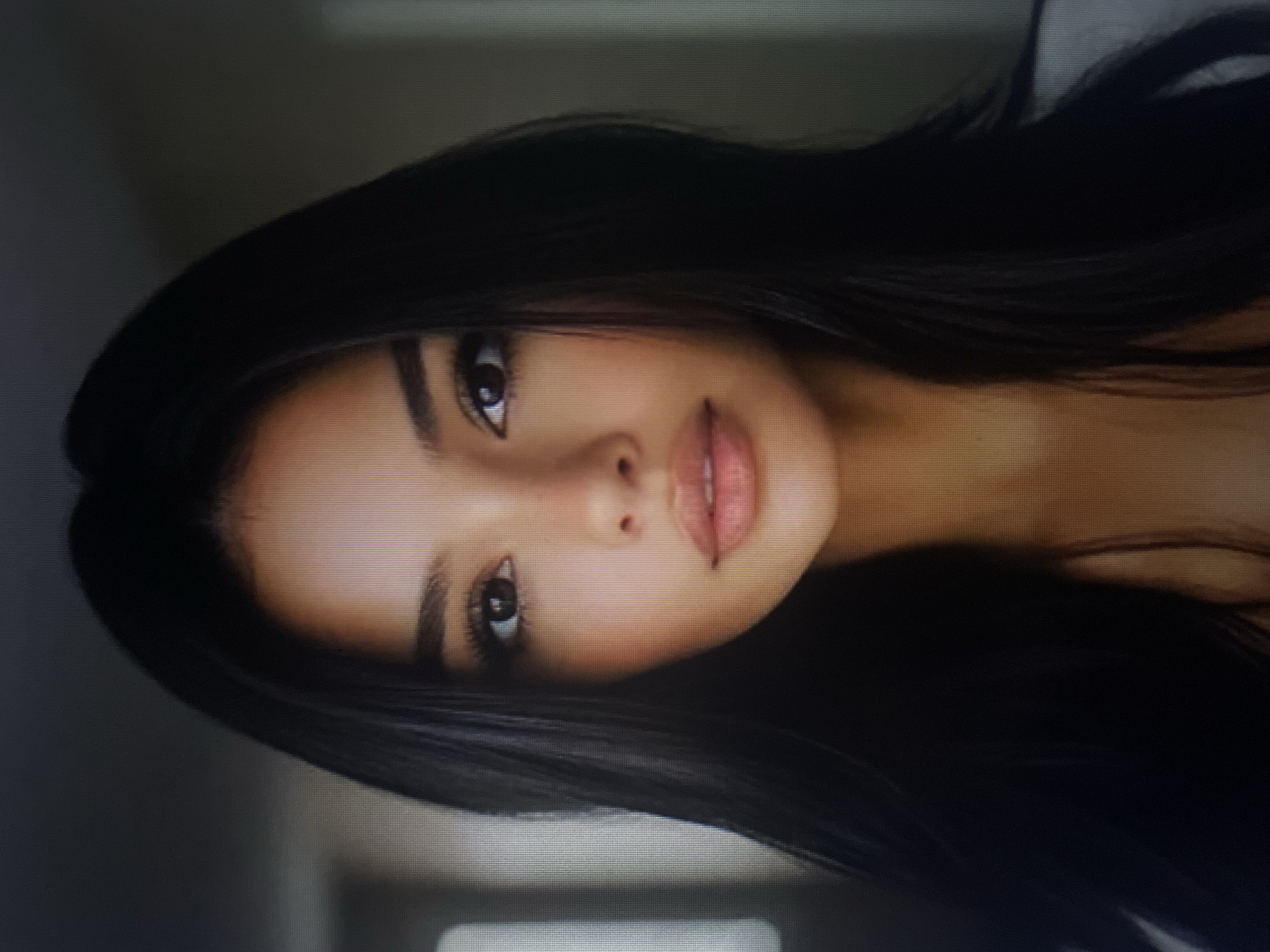 The image is a close-up portrait of a young woman with long, dark hair. She has dark eyes, well-defined eyebrows, and full lips. The background is blurred and appears to be an indoor setting. The woman is looking directly at the camera. There is no information in the image that indicates a location in Kołobrzeg, Poland.