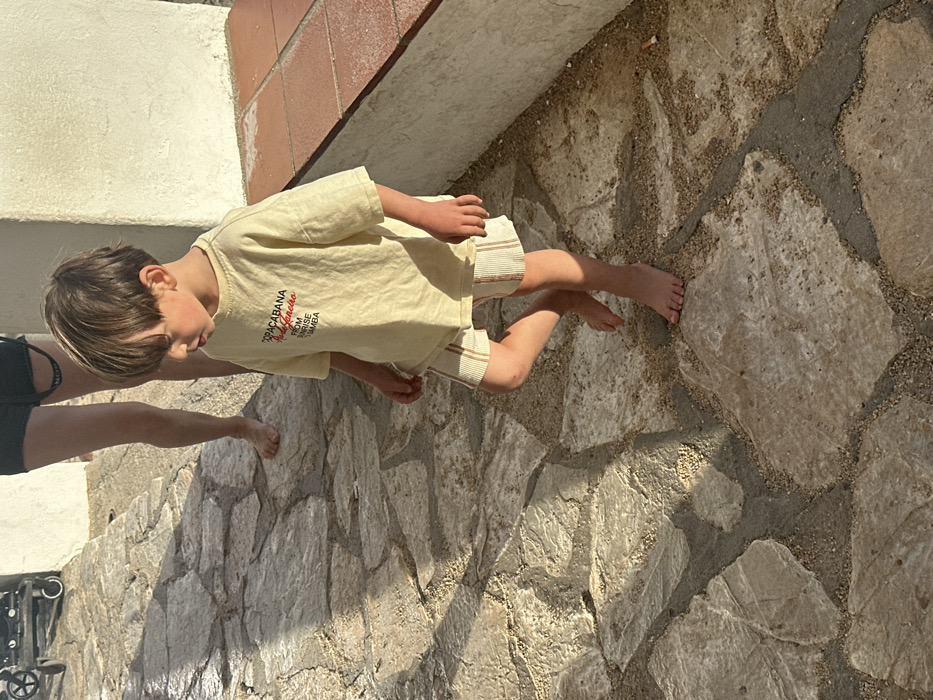 A young child, approximately three to five years old, is depicted in the foreground, walking barefoot on a sunlit, irregularly stone-paved path. The path consists of light-colored flagstones set with visible sand and gravel between them. The child wears a loose-fitting, light-colored (possibly cream or light yellow) t-shirt with red text reading "COPACABANA Rio Janeiro FROM SUNRISE SAMBA" and striped shorts. The child's head is angled downwards and to the left, with brown hair partially obscuring their face. Rosy cheeks are visible. In the background, behind the child, the lower legs and bare feet of an adult, wearing dark shorts, are partially visible. To the right of the child, a light-colored building wall rises, with a terracotta-tiled ledge or step running along its base. Distinct shadows are cast on the ground, indicating bright sunlight. A portion of a black wheeled object, possibly a stroller, is visible in the upper left corner. The scene takes place outdoors in Palafrugell, Spain.