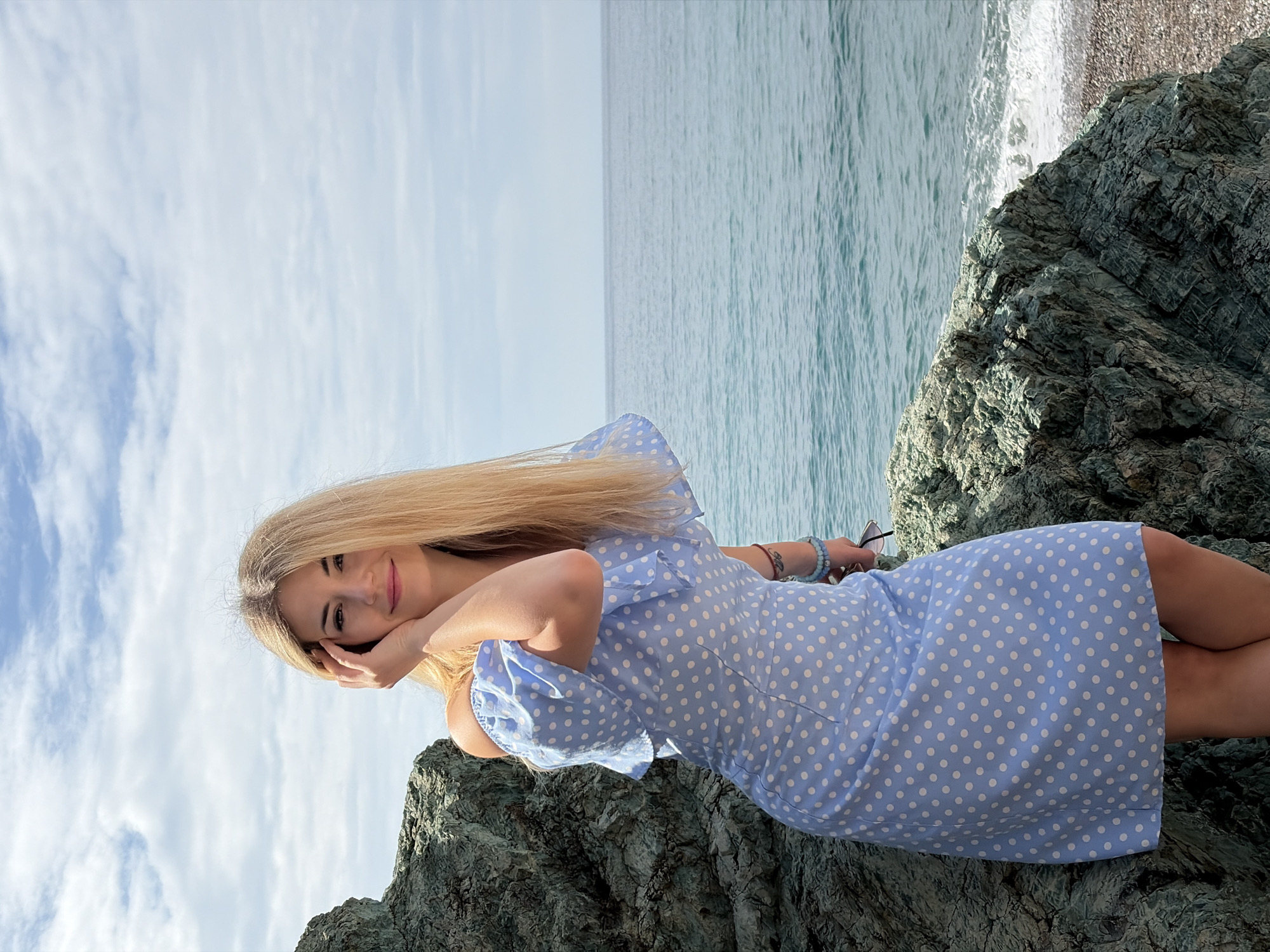 A blonde woman wearing a light blue polka dot dress reclines on a rocky outcrop. She rests her head on her left hand and looks towards the camera. The background reveals a seascape under a cloudy sky. The location is likely Rafailovići, Montenegro, given the coastal setting and rock formations characteristic of the area.