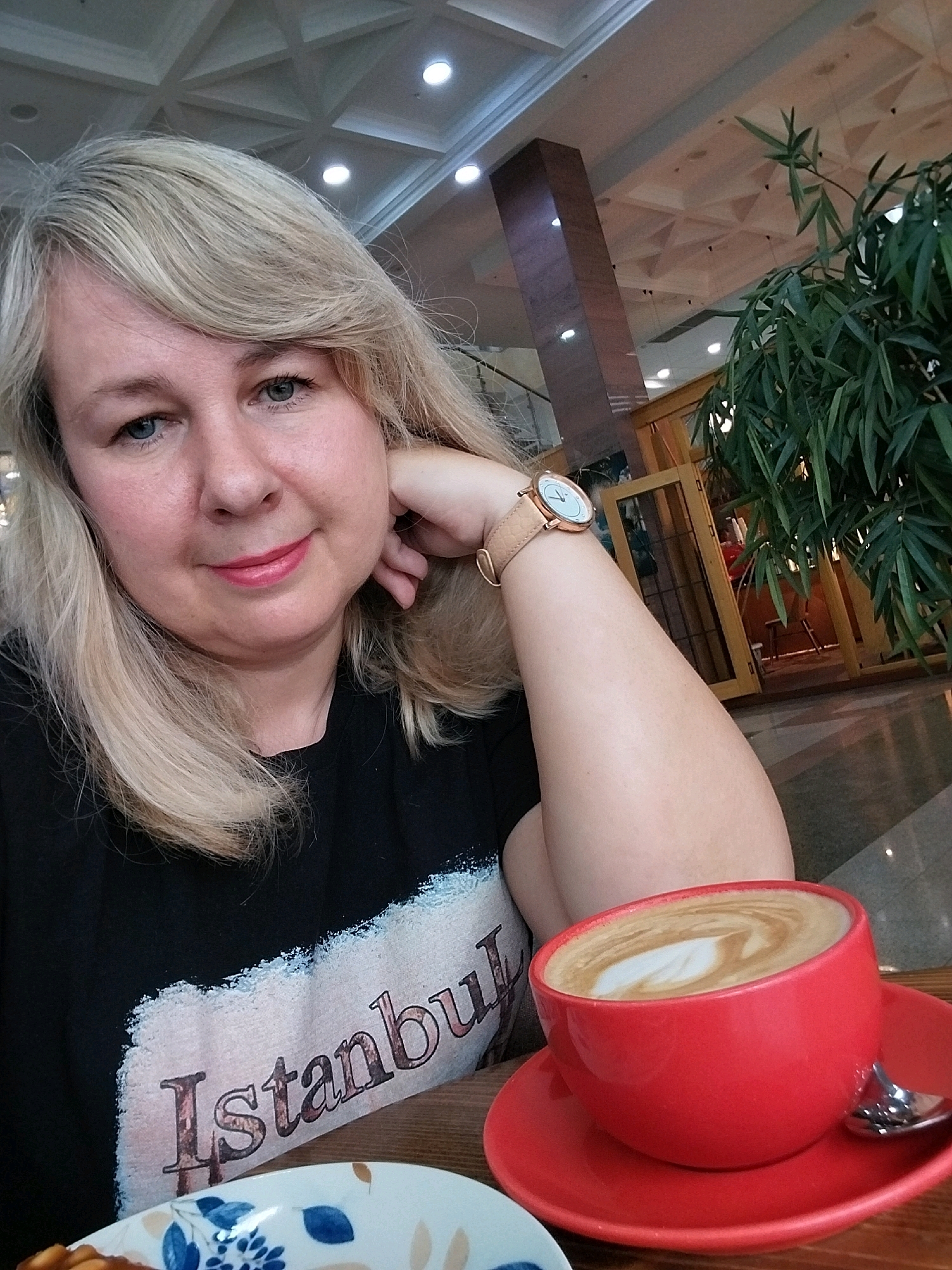 A medium shot photograph captures a woman with blonde hair and blue eyes, resting her chin on her hand. She is wearing a black t-shirt with the word "Istanbul" printed on it. A red cup of coffee with latte art sits on a saucer to her right, accompanied by a spoon. The background suggests an indoor setting, possibly a cafe or restaurant, with a ceiling featuring geometric patterns and lighting, a wooden pillar, and a potted plant. A wooden table is visible in the foreground. The image is taken from a slightly elevated perspective.