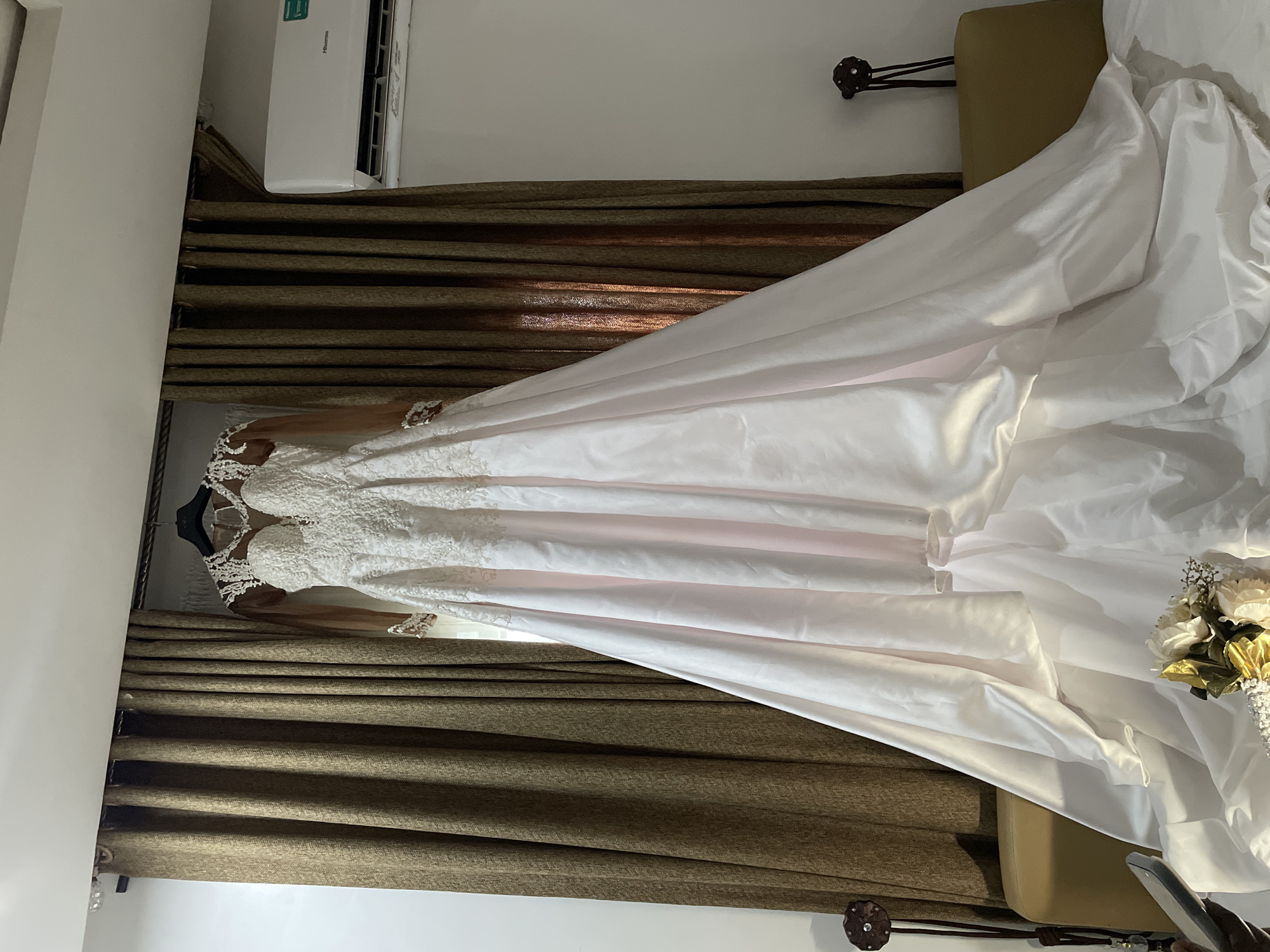 A wedding dress hangs in a room in Jos, Nigeria. The dress is white with lace detailing on the bodice and sleeves. The room has brown curtains and a beige chair. An air conditioner is mounted on the wall.