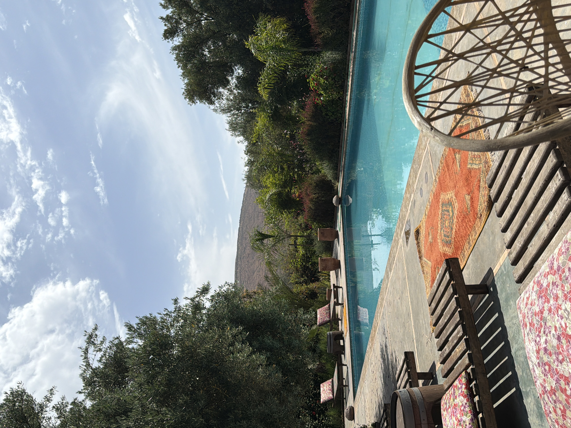 This image depicts a serene outdoor swimming pool area, presented on its side, but intended to be viewed in portrait orientation. When correctly oriented, the scene is dominated by a bright, partly cloudy sky with scattered white cirrus clouds at the top.
The right side of the frame features a clear, light blue swimming pool, reflecting the sky and surrounding foliage. Adjacent to the pool is a paved area where several wooden lounge chairs with colorful, floral-patterned cushions are arranged. A vibrant, ornate rug, primarily in shades of red and orange with intricate patterns, is laid out on the ground near the seating. Strong, angular shadows are cast by the chairs and other poolside furniture, indicating bright sunshine, likely late morning or early afternoon. A large, circular, woven chair back is partially visible in the bottom right corner.
Beyond the pool and its immediate surround, a dense expanse of lush green trees and shrubs forms a natural border, creating a vibrant midground. These include both broadleaf trees and palm-like plants. In the far background, rising majestically above the greenery, is a large, arid mountain range, characteristic of the landscape around Tahannaout, Morocco, under the expansive blue sky. No people or visible text are present in the image. The overall scene evokes a peaceful and relaxing ambiance.