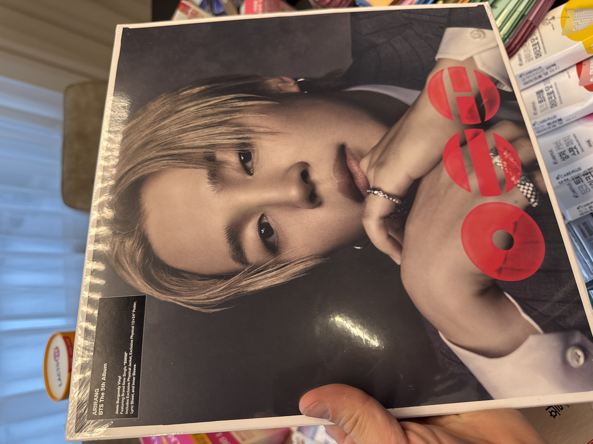 Here is a detailed description of the image:
The image shows a hand holding a vinyl record album of "HOPE" by Jimin of the K-pop group BTS. The album cover features a close-up portrait of Jimin, with the word "HOPE" stylized in red letters. The bottom left corner of the album displays text with "ARIRANG BTS The 5th Album".
In the background, there are visible products with Korean text indicating the location is somewhere in Korea.