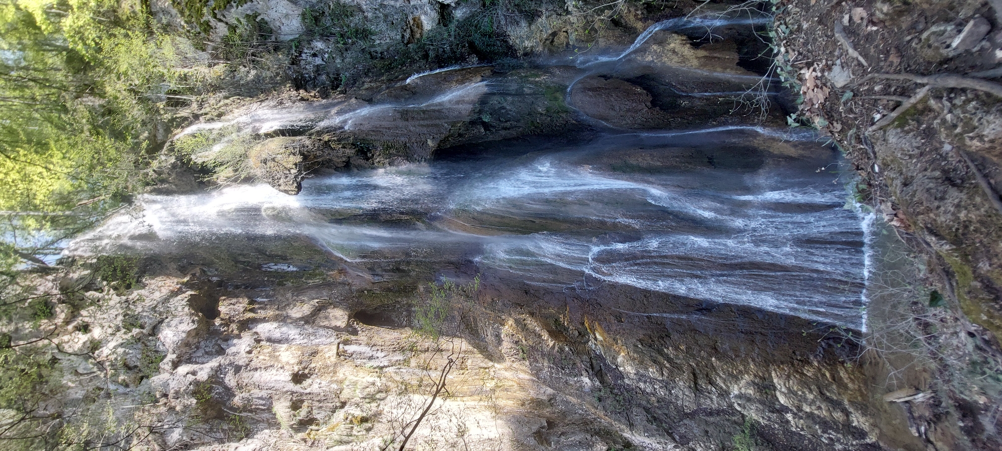 The image shows a waterfall in the area of Бачковски манастир, Bulgaria. Water flows down a rocky cliff face covered with vegetation. The water appears as a blurred white cascade against the darker rocks. The scene is partially shaded, with patches of sunlight hitting the rocks and foliage. The surrounding environment consists of a forest, with trees and undergrowth visible. The image is oriented vertically.