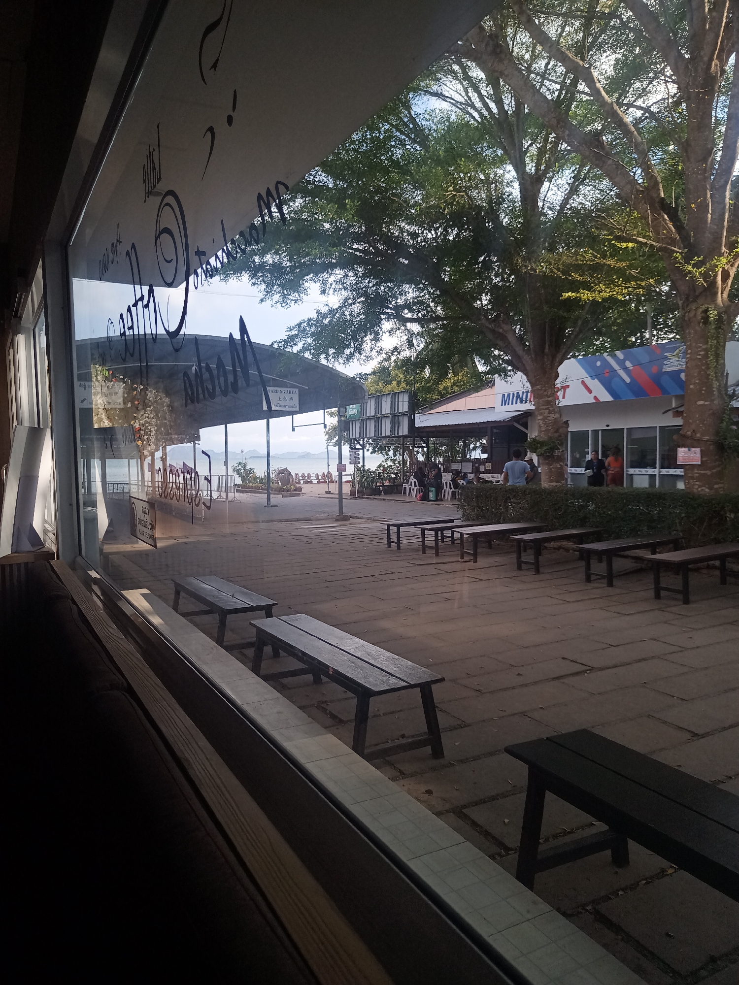 The image shows an outdoor scene in Tambon Hat Sai Ri, Thailand, viewed through a window of a cafe or restaurant. The window has signage displaying "A Little Coffee & More... Mocha". Outside, there is a paved plaza with wooden benches. A "MINIMART" building is visible with a blue, red, and white stripe design, with people standing near its entrance. Further in the background, there is a covered waiting or boarding area, and the sea with distant islands is visible. Trees provide shade to the plaza.