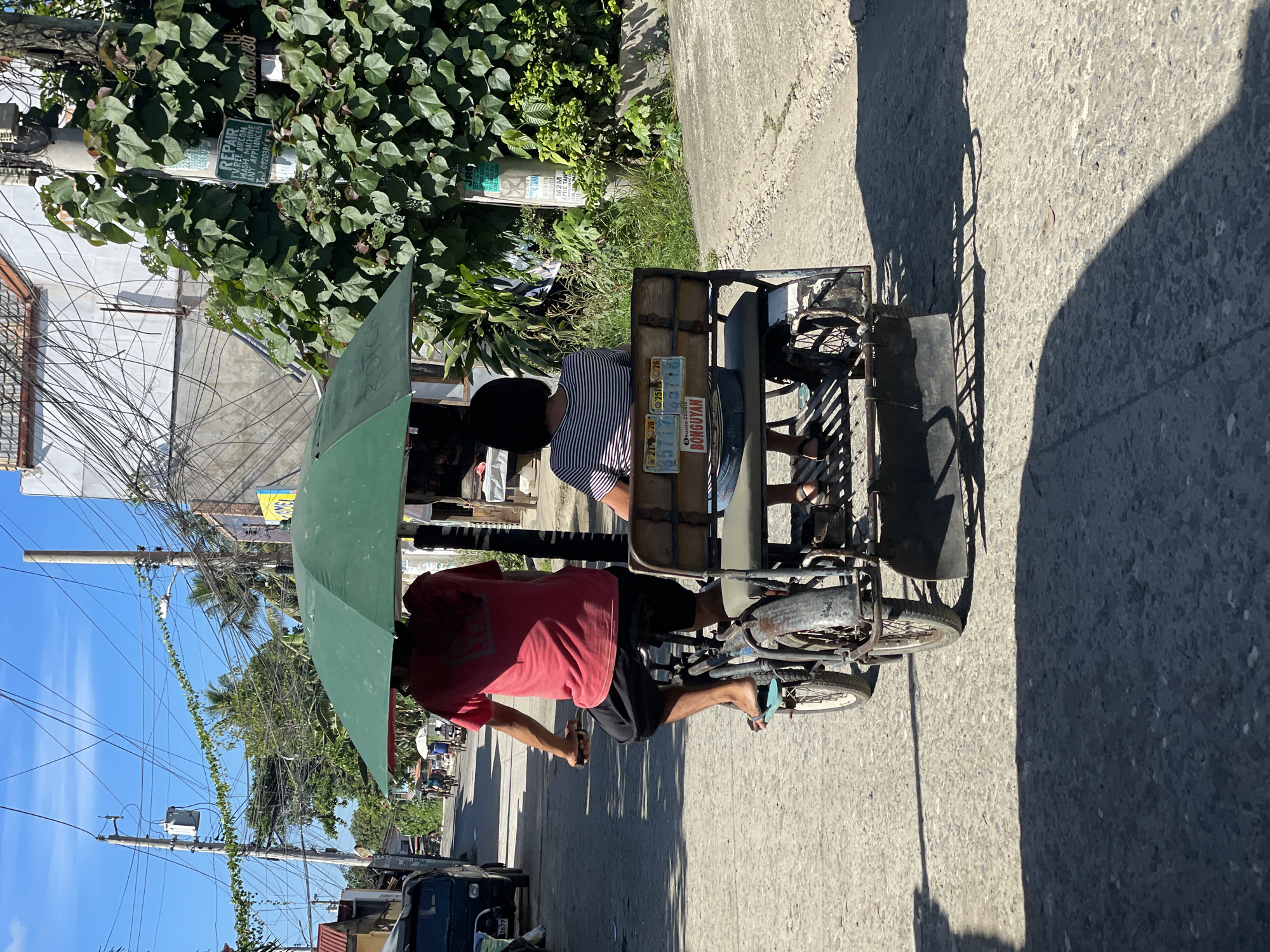 An outdoor, eye-level shot shows a tricycle, a common mode of transport in the Philippines, driving on a paved road in what appears to be a residential area, likely in Davao City, Philippines, given the visual cues such as the architecture and vegetation. The tricycle has a driver in a red t-shirt and black shorts, pedaling while holding a dark green umbrella over the passenger, a woman wearing a striped shirt and shorts. The passenger sits in the back of the tricycle, on a bench seat. The tricycle itself is a modified bicycle with a passenger carriage attached to the side. The road is lined with buildings and dense green foliage, with numerous electrical wires strung overhead, characteristic of many urban and suburban areas in the Philippines. The sunlight creates distinct shadows on the road, indicating a bright day. License plates on the back of the tricycle are visible, with one clearly reading "BONGUYAN". The overall scene captures a slice of daily life and transportation in the Philippines.