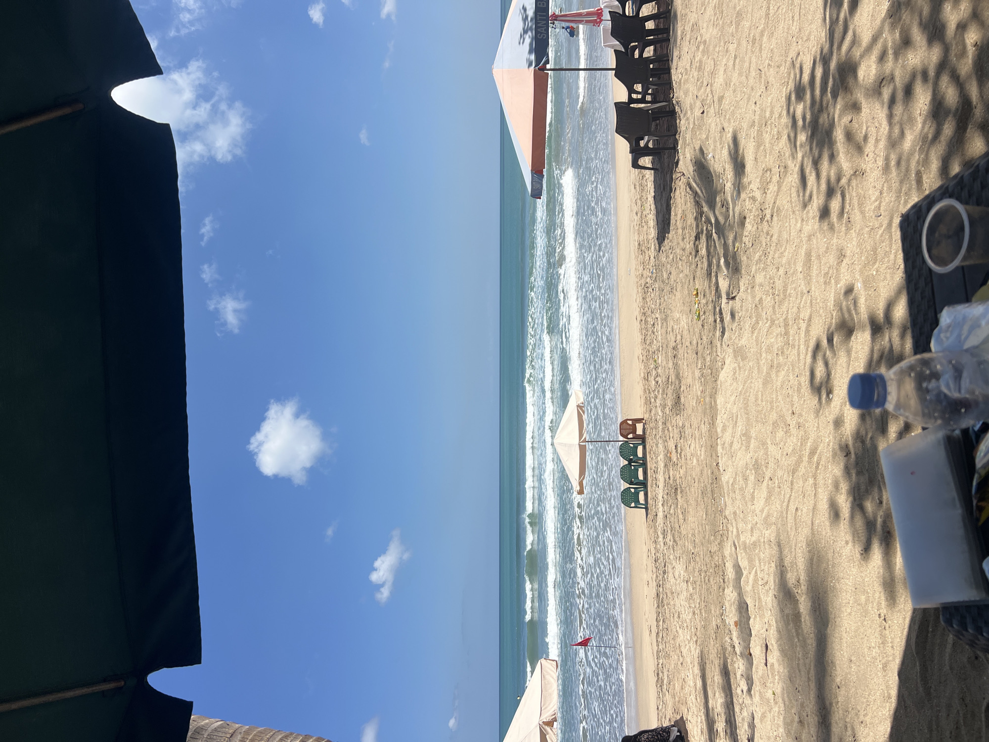 The image captures a scenic view of Kuta Beach in Indonesia on a clear, sunny day. In the foreground, a dark umbrella partially obscures the top of the frame, casting shadows on the sandy beach. A black woven table holds a plastic water bottle, a white object, and a clear plastic cup, suggesting a relaxed setting.
The middle ground features a sandy beach with several beach chairs and umbrellas. Three green chairs and one brown chair are arranged under a small beige umbrella, while a larger, two-toned umbrella with orange and blue panels stands near a row of black plastic chairs. A small red flag is planted on the wet sand near the water's edge.
The background is dominated by the vast turquoise ocean, with gentle waves rolling towards the shore. The horizon line is clear, meeting a bright blue sky dotted with a few fluffy white clouds. The overall impression is one of a tranquil beach day, characteristic of Kuta's popular tourist destination.