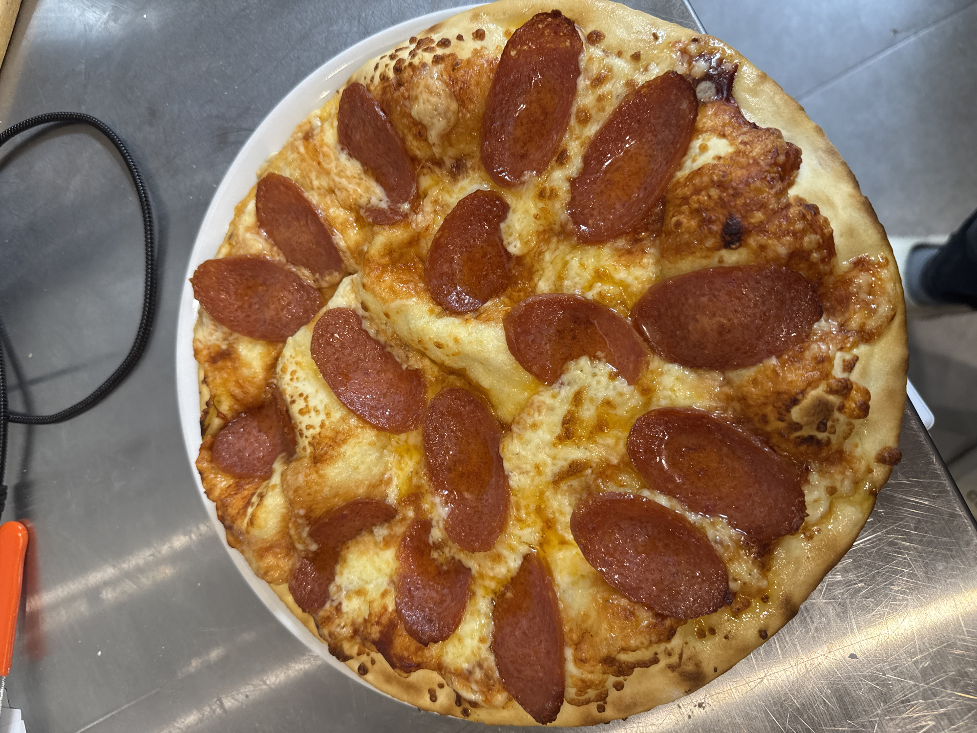A freshly baked pepperoni pizza is the central subject of the image, presented on a round white plate. The pizza is generously topped with numerous circular slices of pepperoni and melted, golden-brown cheese, showing some bubbly and slightly browned spots from baking. The crust is well-cooked, with a golden-brown hue and some darker, crispier areas.
The pizza and plate rest on a metallic, possibly stainless steel, surface, which appears to be a counter or table. This surface has visible scuffs and reflections, suggesting an indoor setting like a kitchen or commercial food preparation area. In the top-left background, a coiled black cable is visible. To the bottom-left, a small portion of an orange handle, possibly from a knife or tool, peeks into the frame. Faintly visible in the bottom right, a person's shoe can be discerned, indicating human presence near the pizza.
The image captures the pizza as if it has just been served or is about to be eaten. There is no discernible activity or event beyond the presentation of the food. No text is visible. The time of day and weather cannot be determined, but the lighting suggests an indoor environment. The location context provided is Yerevan, Armenia, though no specific visual cues in the image confirm this.