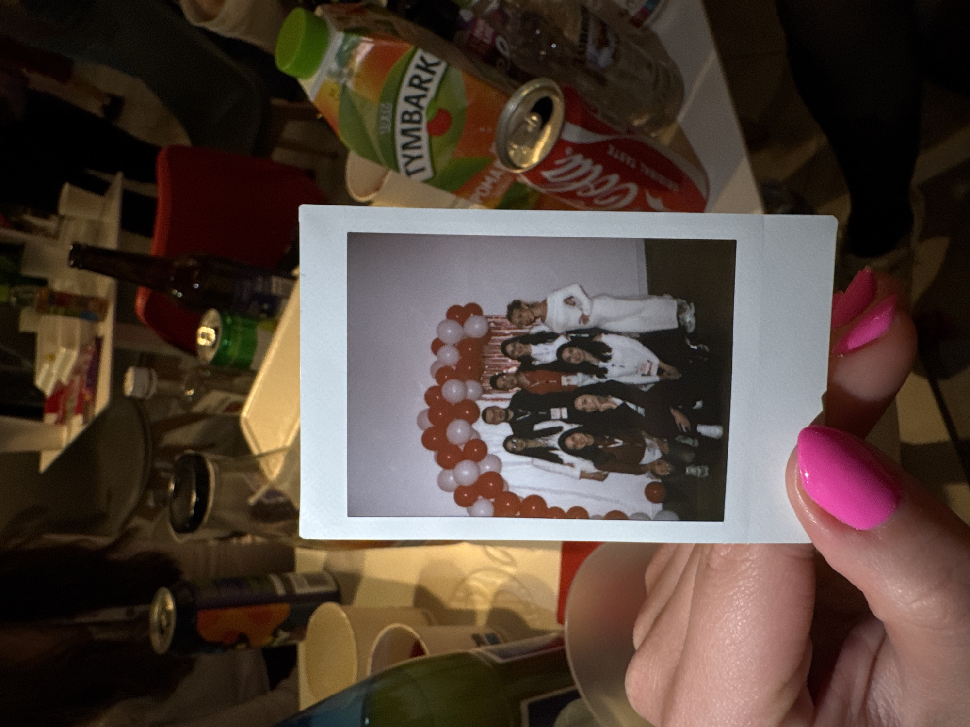 The image shows a Polaroid photo being held in a hand with pink nail polish. The Polaroid depicts a group of people lying on the floor surrounded by red and white balloons. The individuals are dressed in a mix of white and dark clothing.
The photo is being held over a cluttered surface with various objects including a juice box (Tymbark), a can of Coca-Cola, and various other bottles and containers. The setting appears to be indoors. There are no elements to indicate the location to be in the city of Bydgoszcz, Poland.