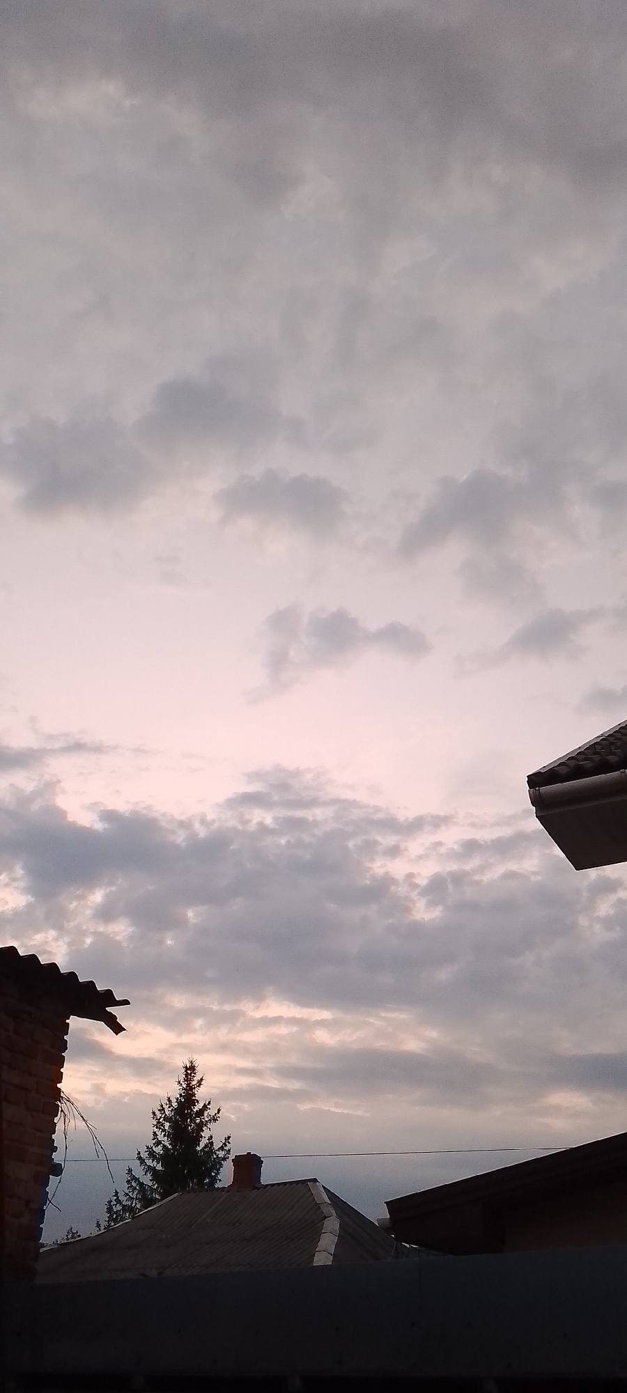 The image depicts a cloudy sky over rooftops, likely in Belgorod, Russia, given the context. The sky is filled with various shades of gray and pale pink clouds, suggesting either dawn or dusk. In the foreground, the roofs of several buildings are visible. On the left, a portion of a brick wall and a dark-tiled roof are present. Further back, a corrugated metal roof is visible with a chimney. A tall, dark green coniferous tree stands behind this roof. To the right, a modern-looking dark roof with a gutter system is angled into the frame. The overall scene is quiet and appears to be an outdoor view from an elevated position, possibly a window or balcony. There are no people or animals visible in the image.