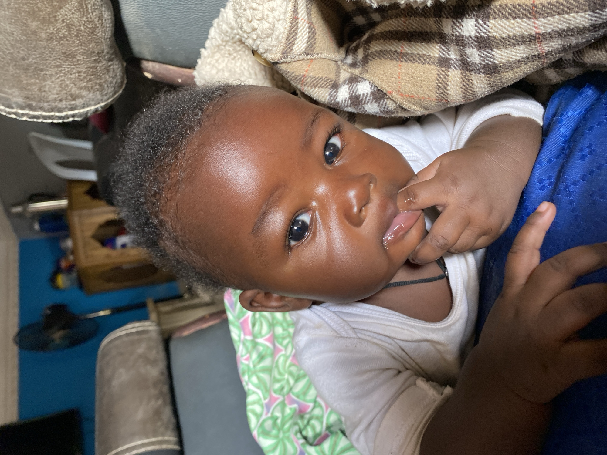 A close-up, slightly tilted portrait captures an alert infant with dark skin and short, dark hair, indoors in what is presumably Kuru Karama, Nigeria. The baby, wearing a white top, looks directly at the camera with wide, curious eyes. Their right hand is positioned near their mouth, with fingers possibly being sucked on, indicated by visible saliva.
The baby is situated amongst various soft furnishings. A plaid blanket, featuring beige, brown, and white squares, is visible behind their head and shoulders, alongside a cream-colored, textured fabric. Below the baby, a green and pink patterned fabric is partially seen, while a royal blue fabric appears to the baby's side, possibly part of clothing or another blanket.
The background is softly blurred but reveals a blue wall, a wooden cabinet or shelf, and a dark floor fan, suggesting a typical domestic interior. The lighting is soft and consistent, indicative of an indoor setting during daylight hours. The infant's expression is one of engagement and curiosity. There is no visible text in the image.
