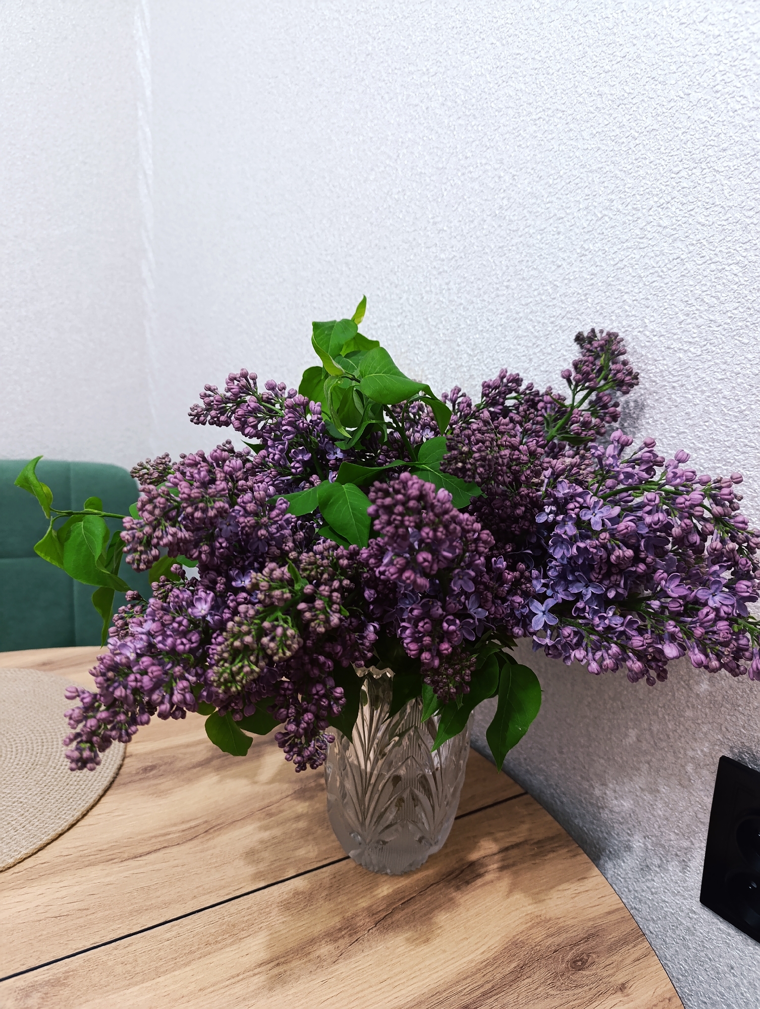 A still-life photograph depicts a bouquet of purple lilacs arranged in a clear, faceted glass vase. The vase sits on a light brown, wood-grain-patterned round table. A tan placemat is visible at the left edge of the table. Behind the bouquet, a textured white wall provides a backdrop. To the left of the flowers, a glimpse of a dark green upholstered object, possibly a chair or sofa, is visible. A black electrical outlet is affixed to the wall on the right side of the frame. The scene is indoors, and the lighting appears to be soft and diffused. The location is unknown.