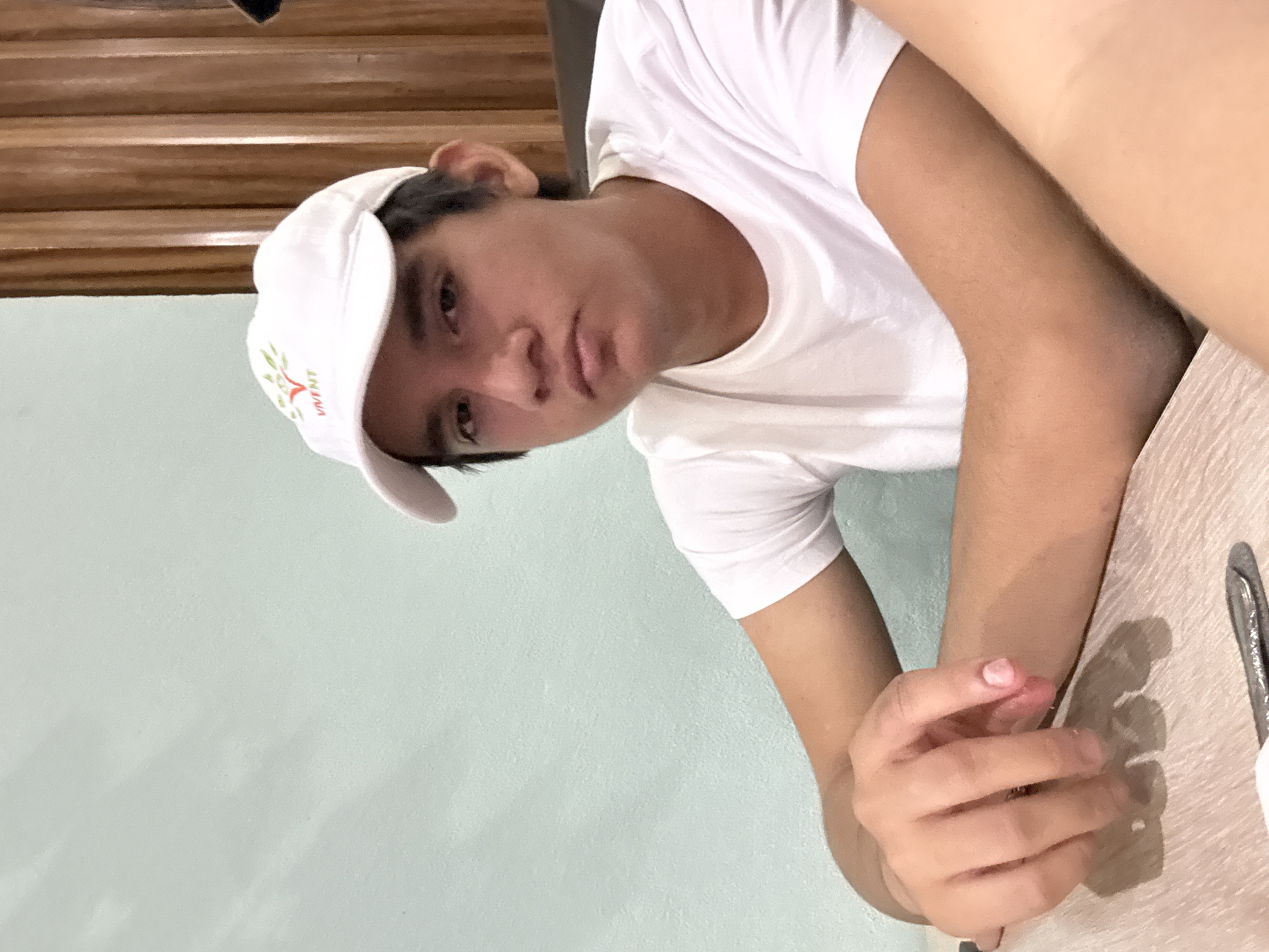 A young man wearing a white t-shirt and a white baseball cap with a green logo sits at a table in front of a light blue wall. He appears to be leaning on the table with his left arm and is looking directly at the camera with a neutral expression. A partially visible wooden structure is behind the wall in the background. The image's location in San Bernardino Tlaxcalancingo, Mexico is not apparent.