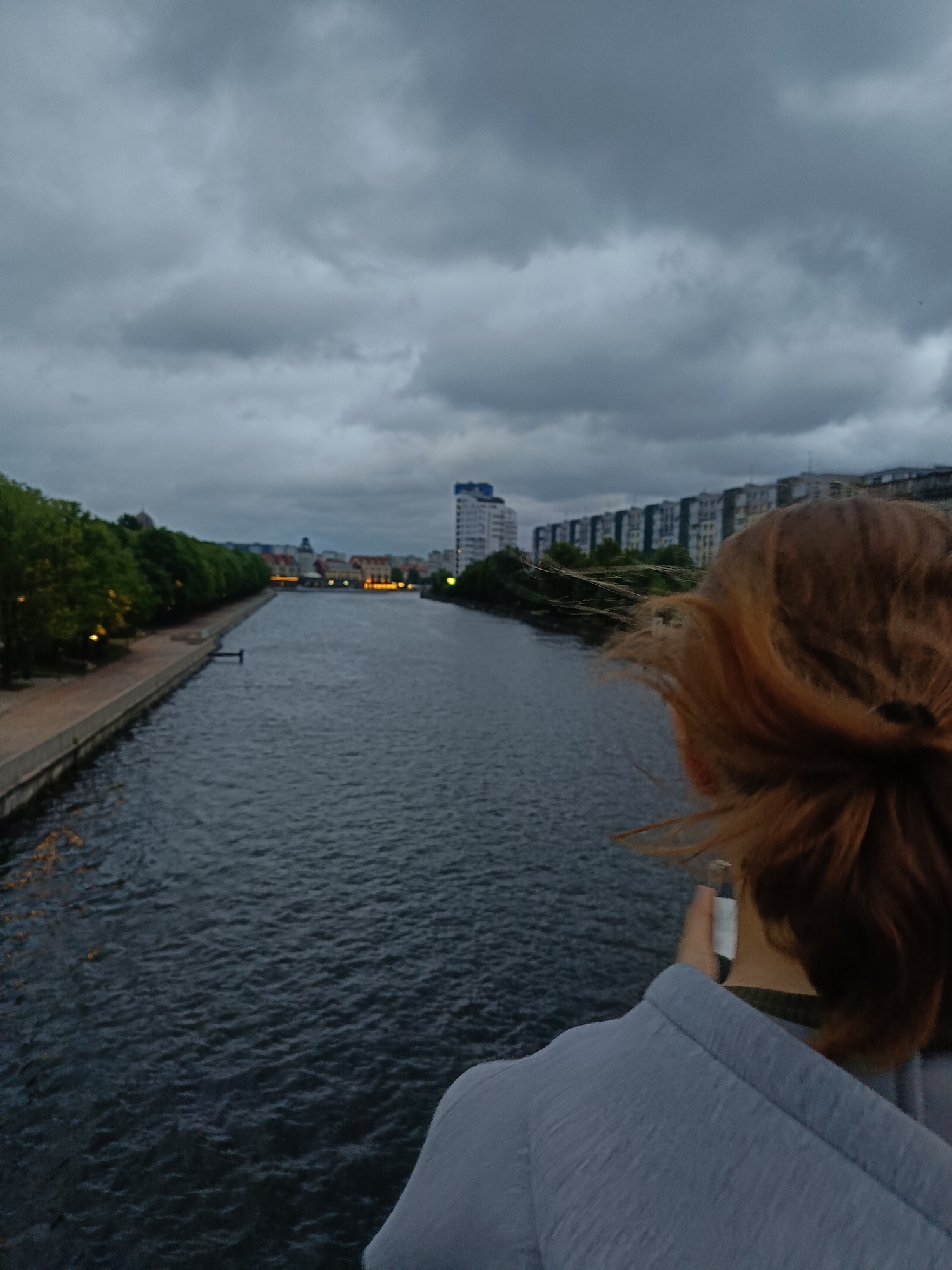 The image shows a view from a bridge in Kaliningrad, Russia, looking out over the Pregolya River. In the foreground, the back of a person's head and shoulders are visible, with their light brown hair blowing in the wind. The person appears to be wearing a light blue hooded jacket. The river flows away from the viewer, with a paved walkway and trees lining the left bank. In the distance, buildings and a taller structure are visible on the right bank, and lights can be seen reflecting on the water. The sky is overcast and cloudy, suggesting it is either early morning or late evening. The overall atmosphere is somewhat moody due to the dark clouds and muted lighting.